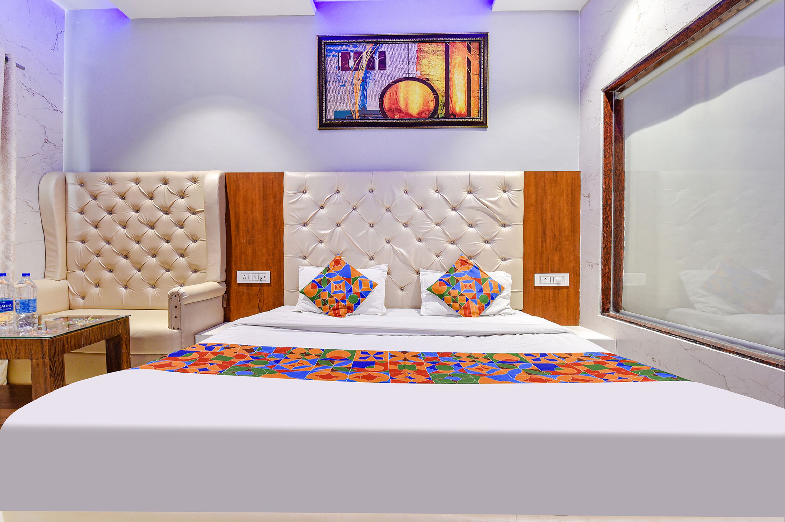 A hotel room with a tufted headboard, two pillows, a colorful bedspread, a wooden side table, and a painting on the wall.