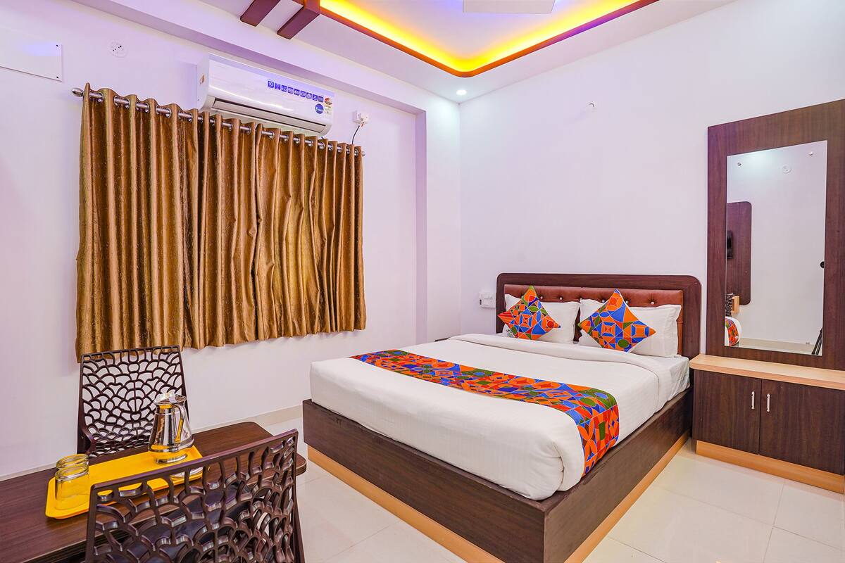 A hotel room with a bed, a desk, a chair, a mirror, and an air conditioning unit.