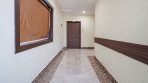 Un couloir avec une porte en bois au fond, un sol carrelé et une fenêtre avec des stores.
