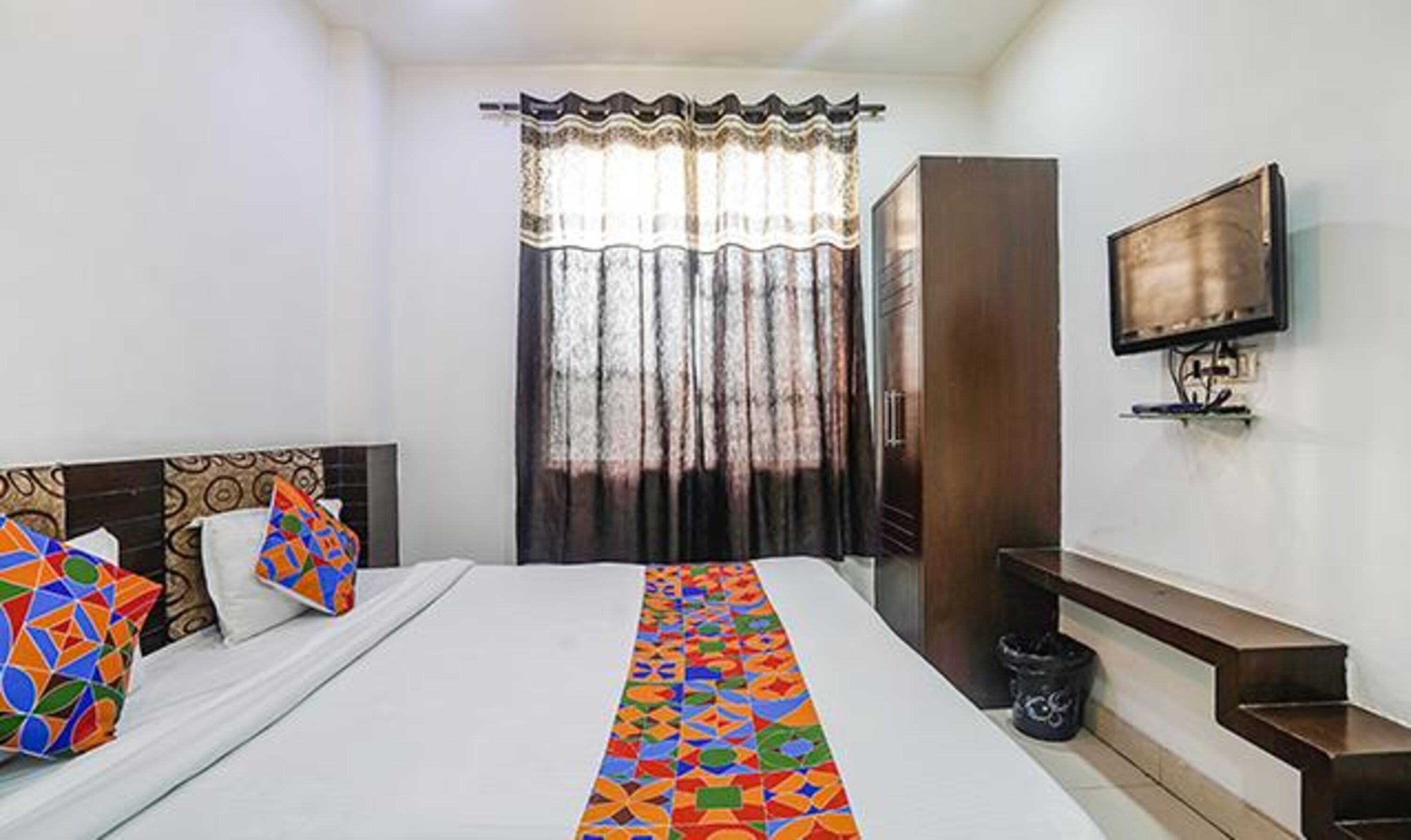 A hotel room with a bed, a TV mounted on the wall, a wardrobe, and a window with curtains.