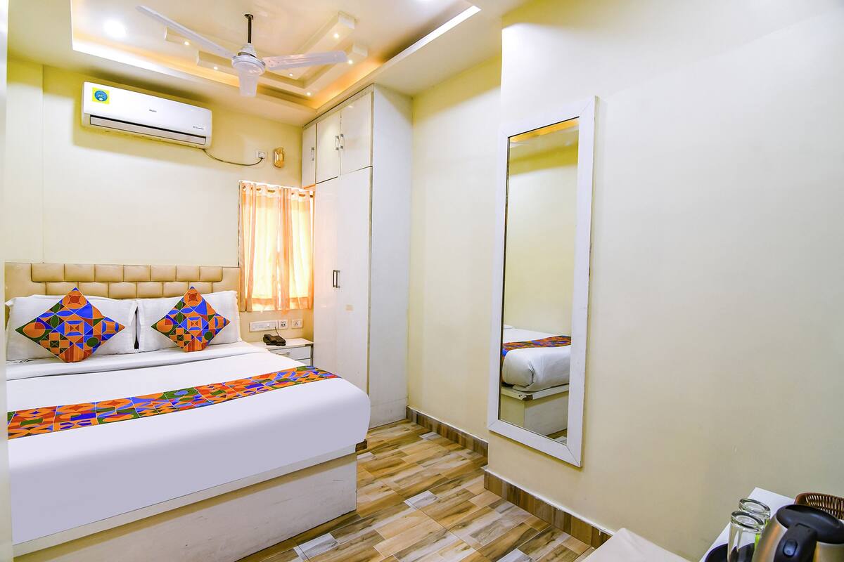 A hotel room with a bed, a ceiling fan, an air conditioning unit, a mirror, and a wooden floor.