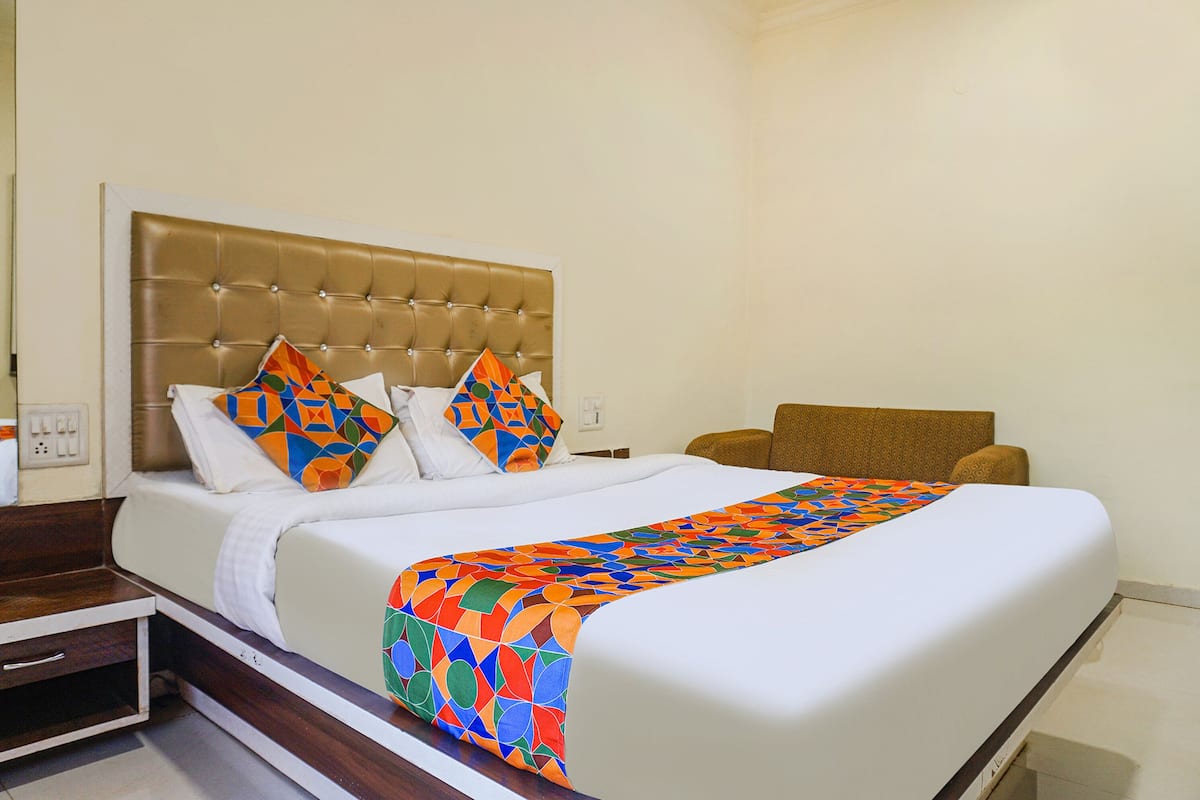 Habitación de hotel con una cama grande, cabecera tapizada en marrón, ropa de cama blanca y almohadas y una colcha con estampados geométricos coloridos.