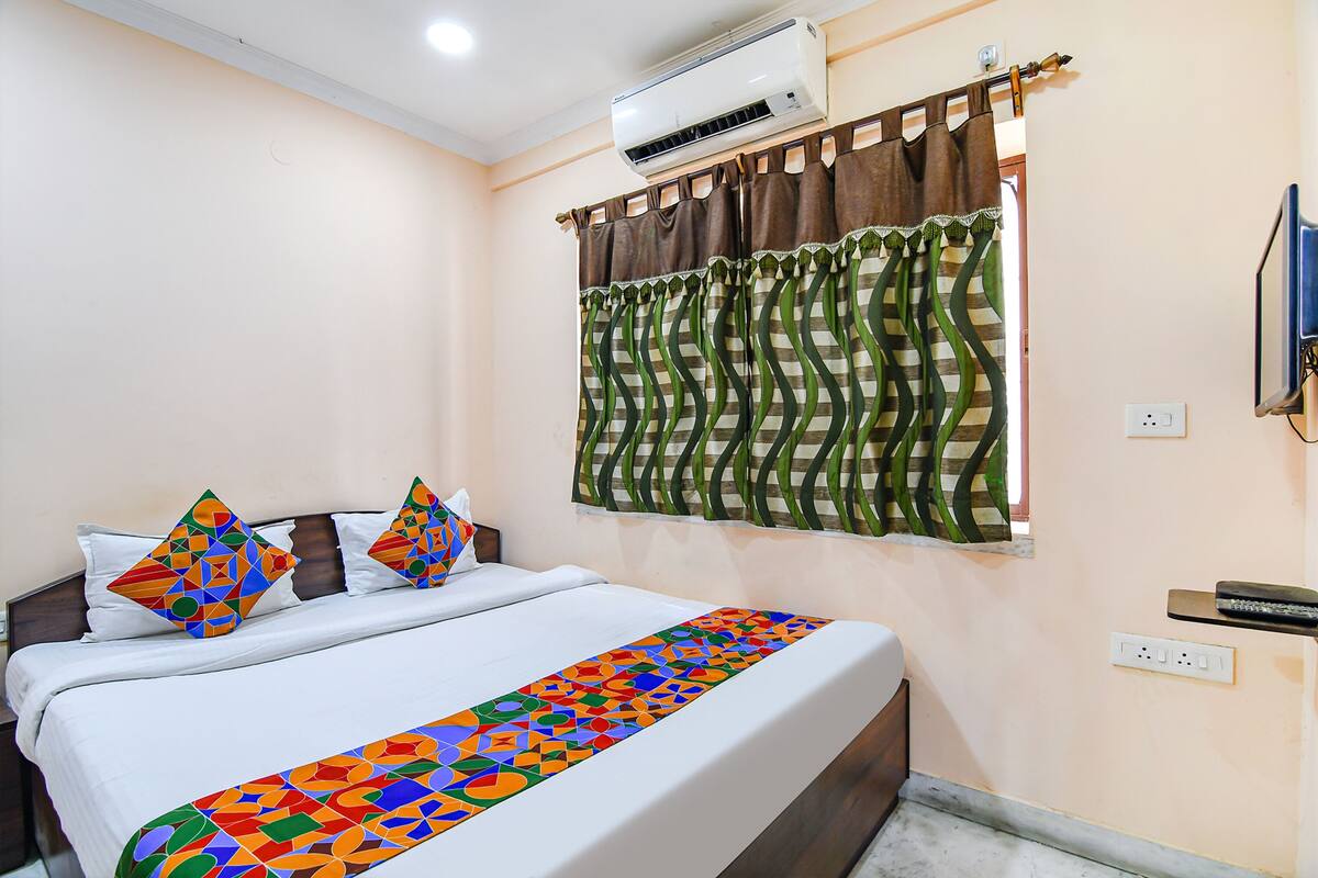 A hotel room with a bed, air conditioning unit, window with curtains, and a television.