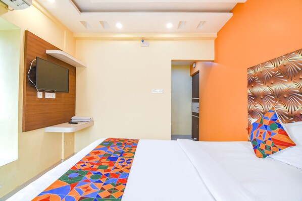 A hotel room with a bed, a TV mounted on the wall, a small shelf, and an orange accent wall.