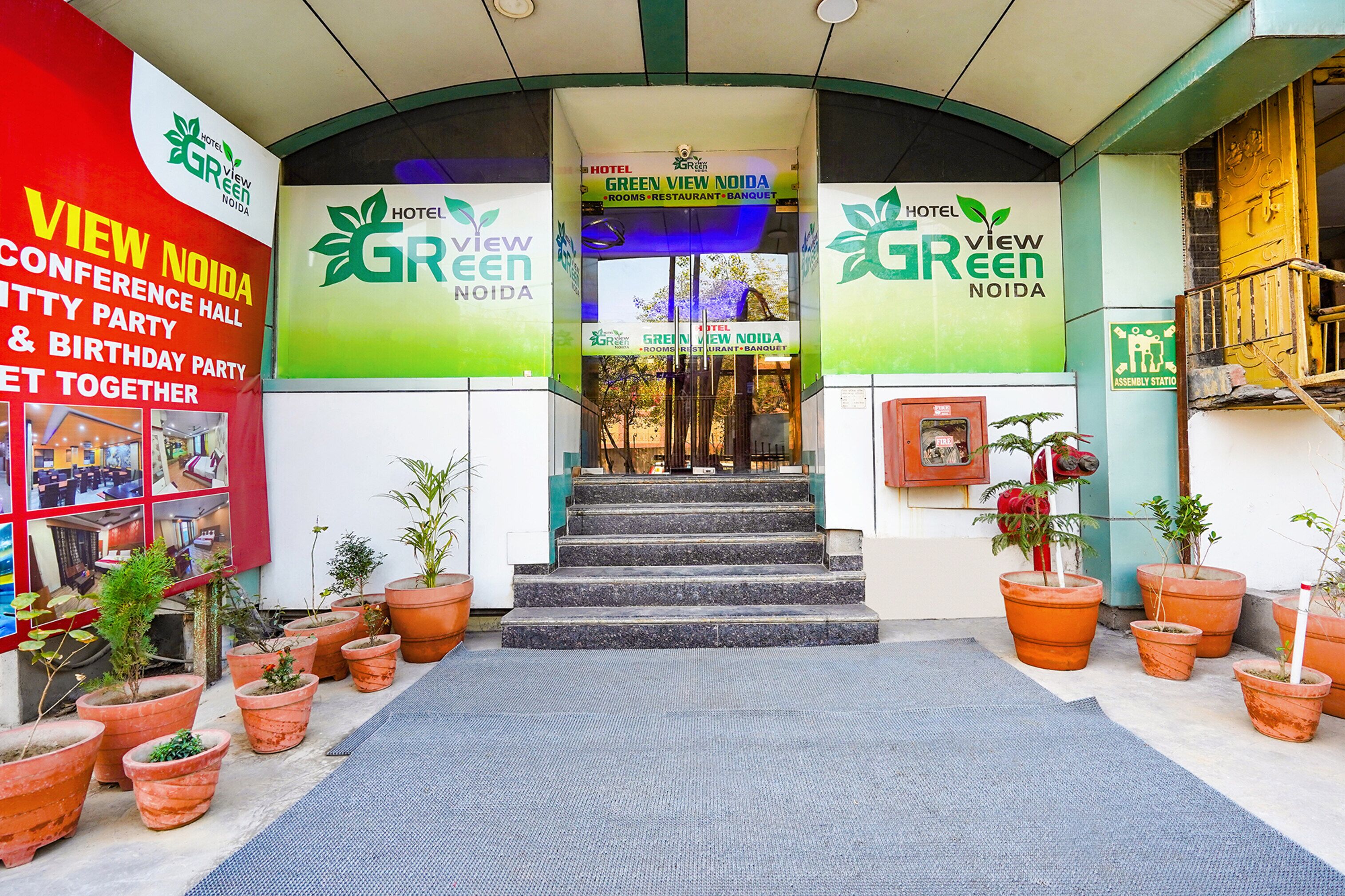 Entrada do hotel Green View Noida, com uma escadaria que leva à entrada, cercada por plantas em vasos.