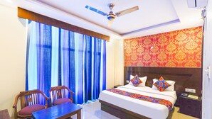 Quarto de hotel com cama, escrivaninha, duas cadeiras, ventilador de teto e janela com cortinas azuis.