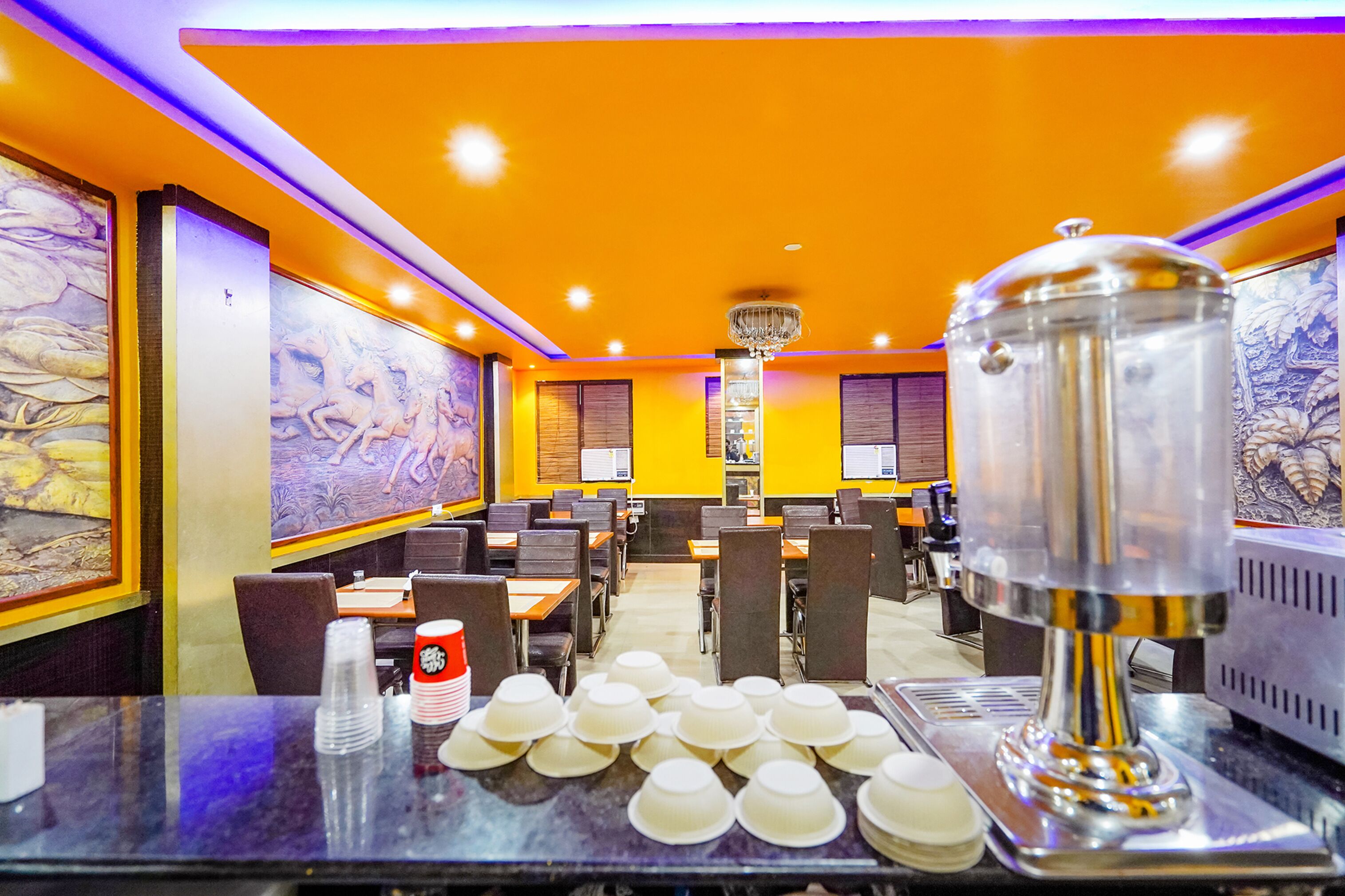 Un restaurant aux murs orange, avec de nombreuses tables et des chaises. Les murs sont décorés de tableaux et un grand distributeur d’eau est posé sur le comptoir.