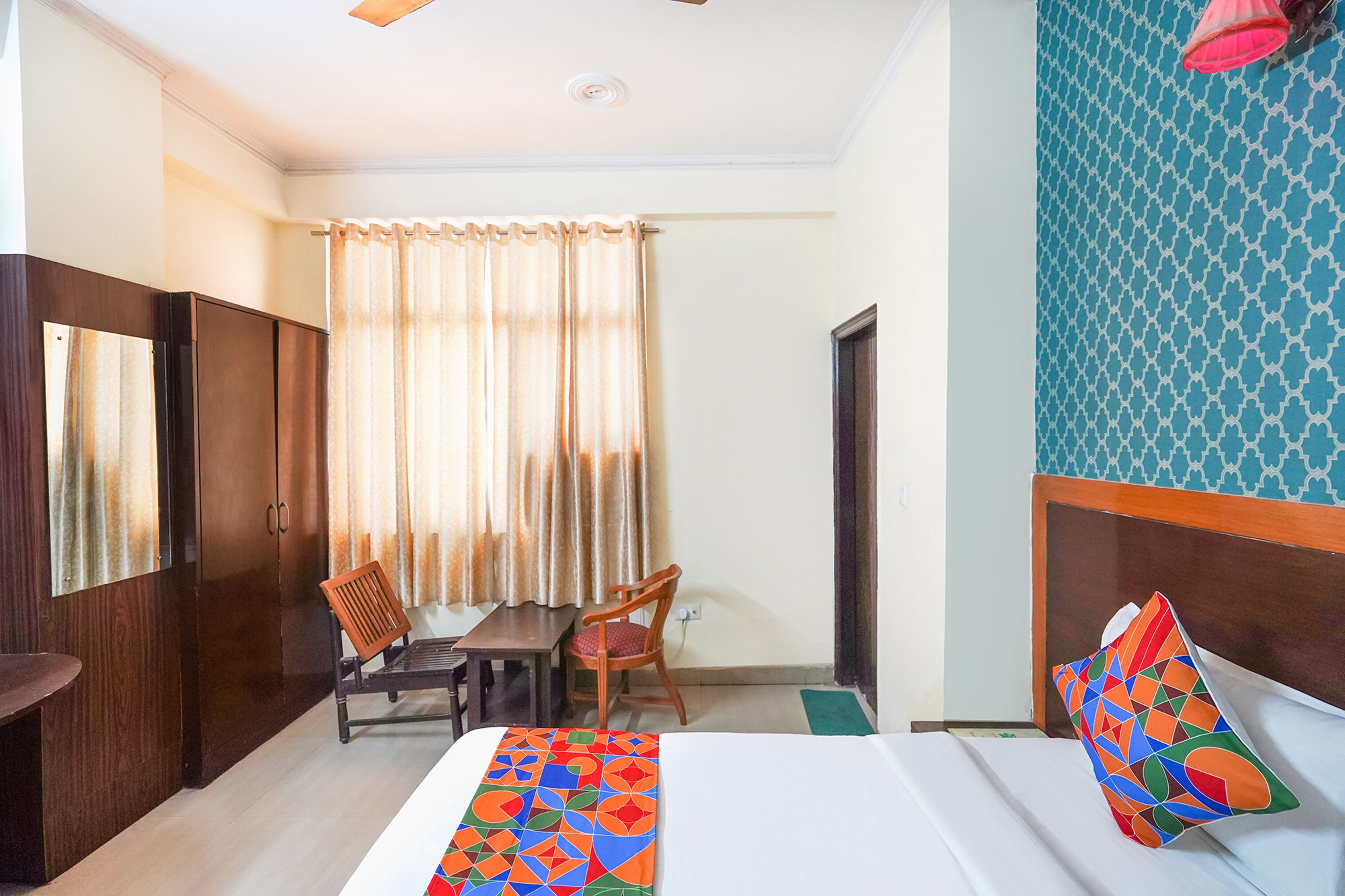 Une chambre d’hôtel avec un lit, une armoire en bois, une table avec une chaise, une fenêtre avec des rideaux et un mur à motifs.