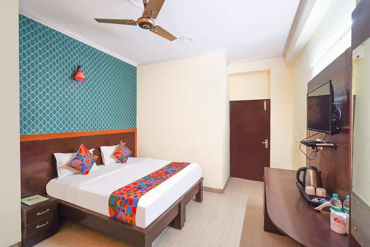 A hotel room with a bed, a nightstand, a desk with a kettle, a TV, and a ceiling fan.