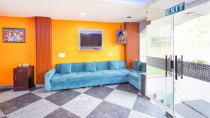 Um saguão de hotel com um sofá azul, uma placa de saída, uma televisão e um quadro na parede.