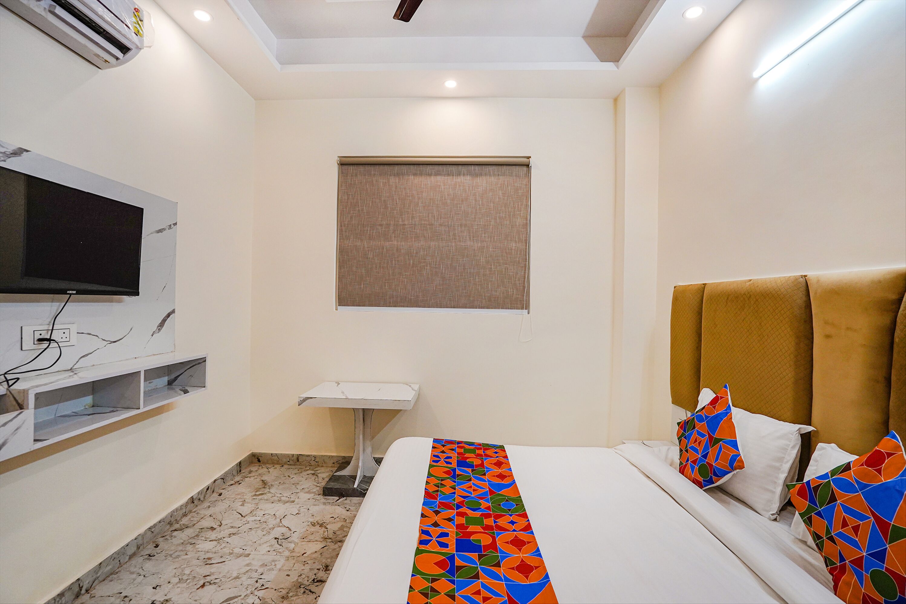 A hotel room with a bed, a TV mounted on the wall, a small table, and a window with a blind.
