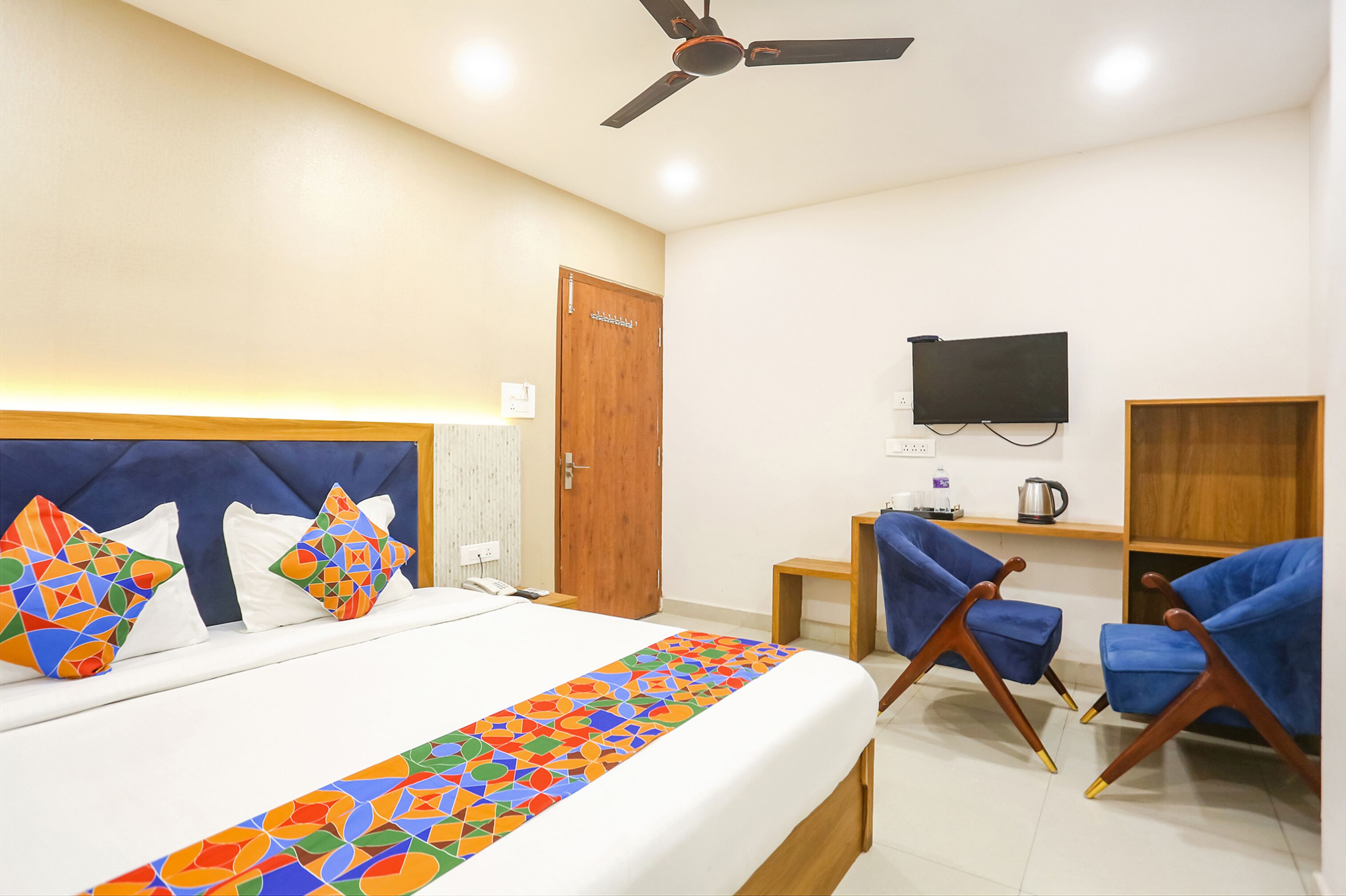 Habitación de hotel con cama, ventilador de techo, televisor montado en la pared y dos sillas azules.
