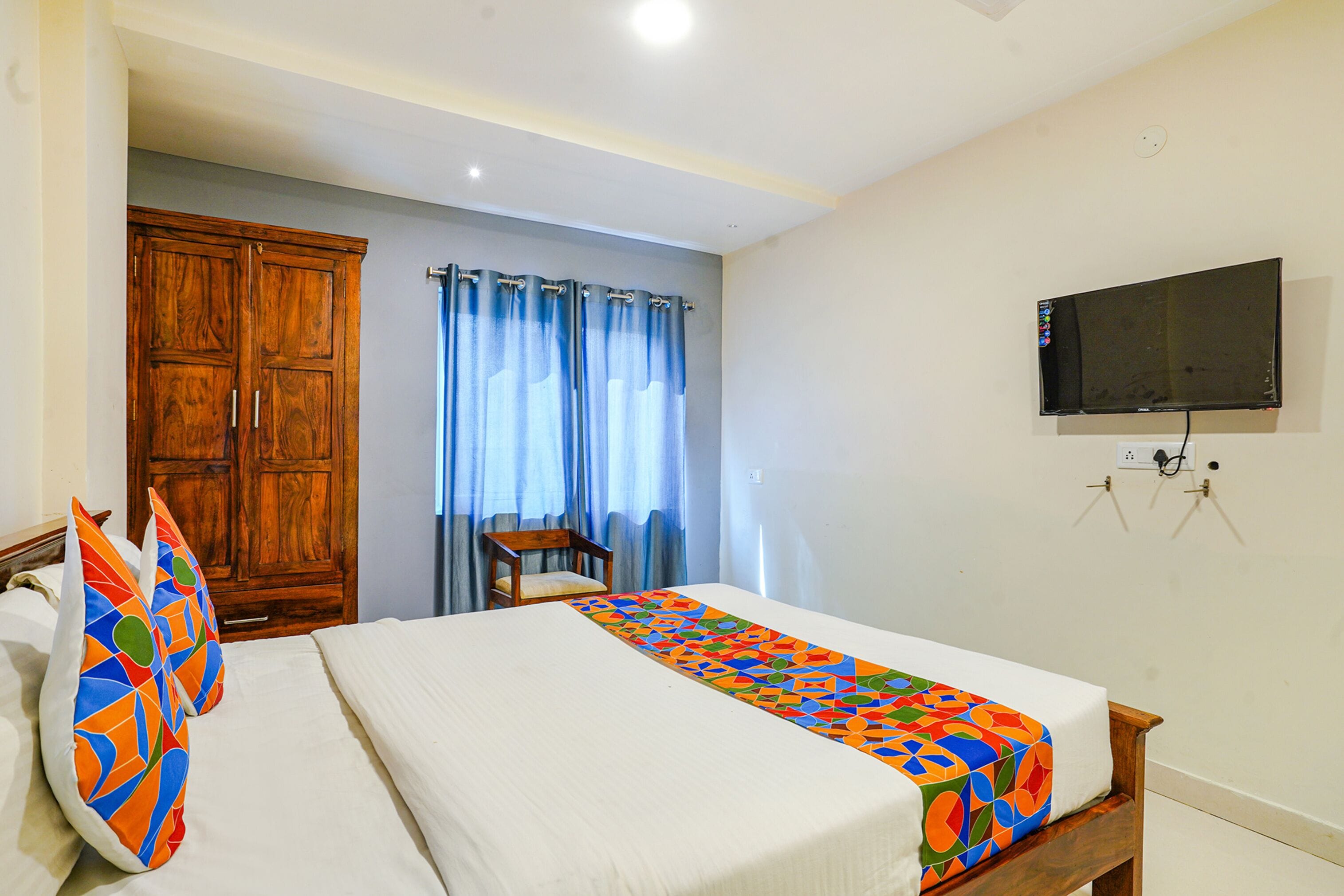 A hotel room with a bed, a wooden wardrobe, a chair, a television mounted on the wall, and a window with curtains.