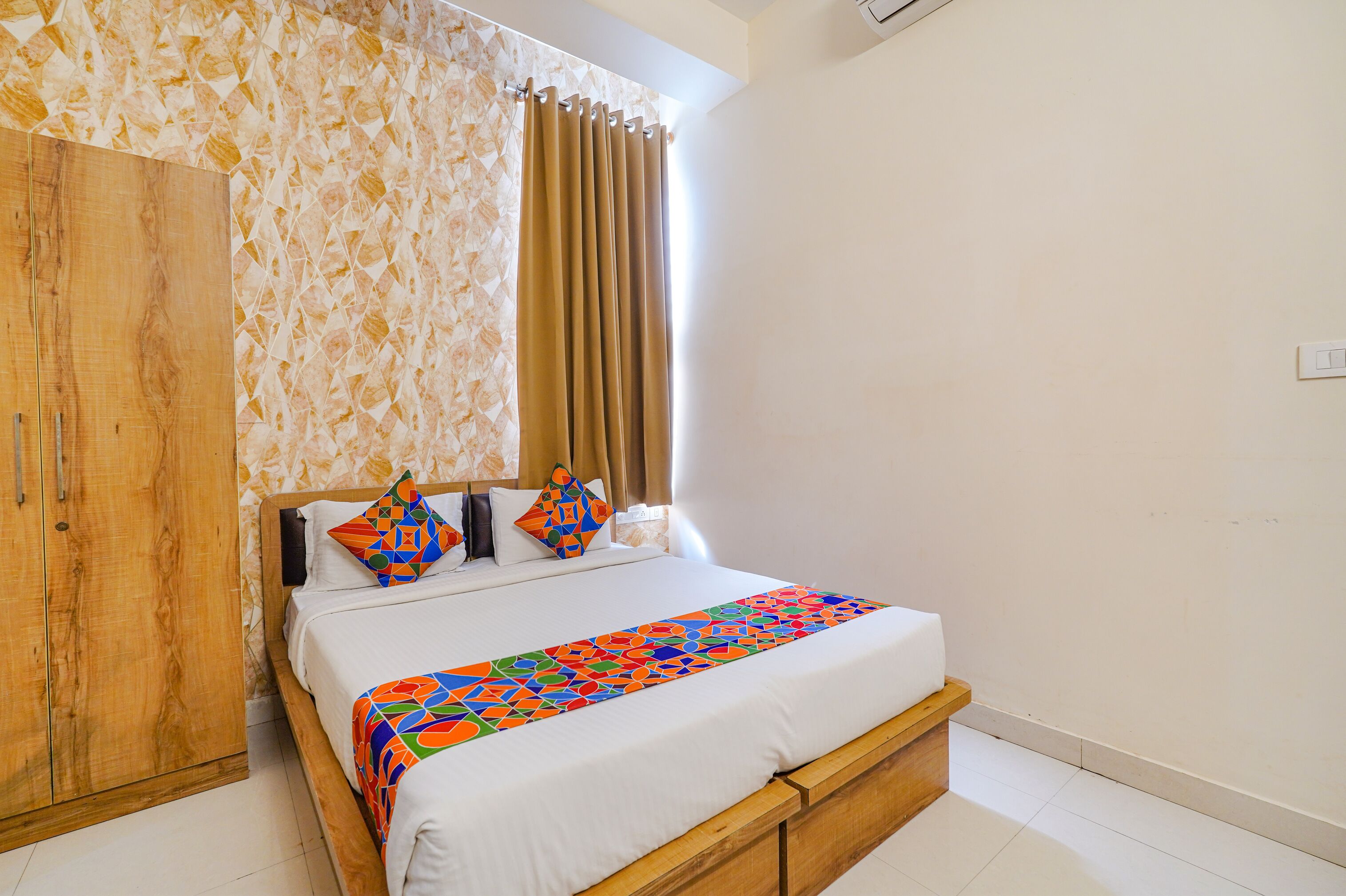 A neatly made bed with a colorful geometric-patterned bedspread, a wooden wardrobe, and a patterned wall.