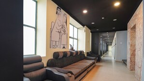 Um hall de hotel moderno com sofás de couro, um grande mural abstrato e um corredor longo com portas.