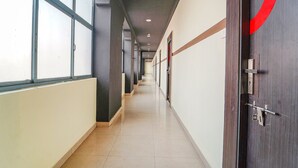 Um corredor comprido e estreito, com chão de azulejos, grandes janelas de um lado e uma porta de madeira com um desenho circular vermelho do outro.
