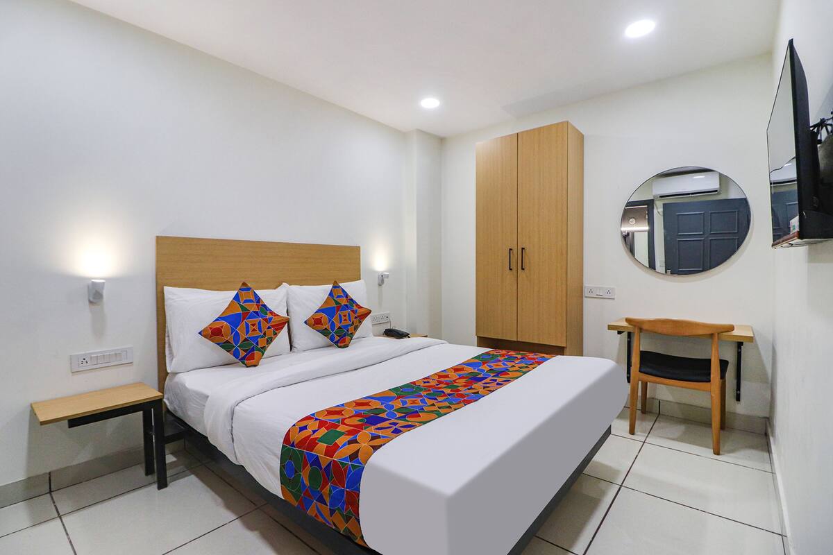 A hotel room with a bed, a wooden wardrobe, a small table, a chair, and a TV mounted on the wall.