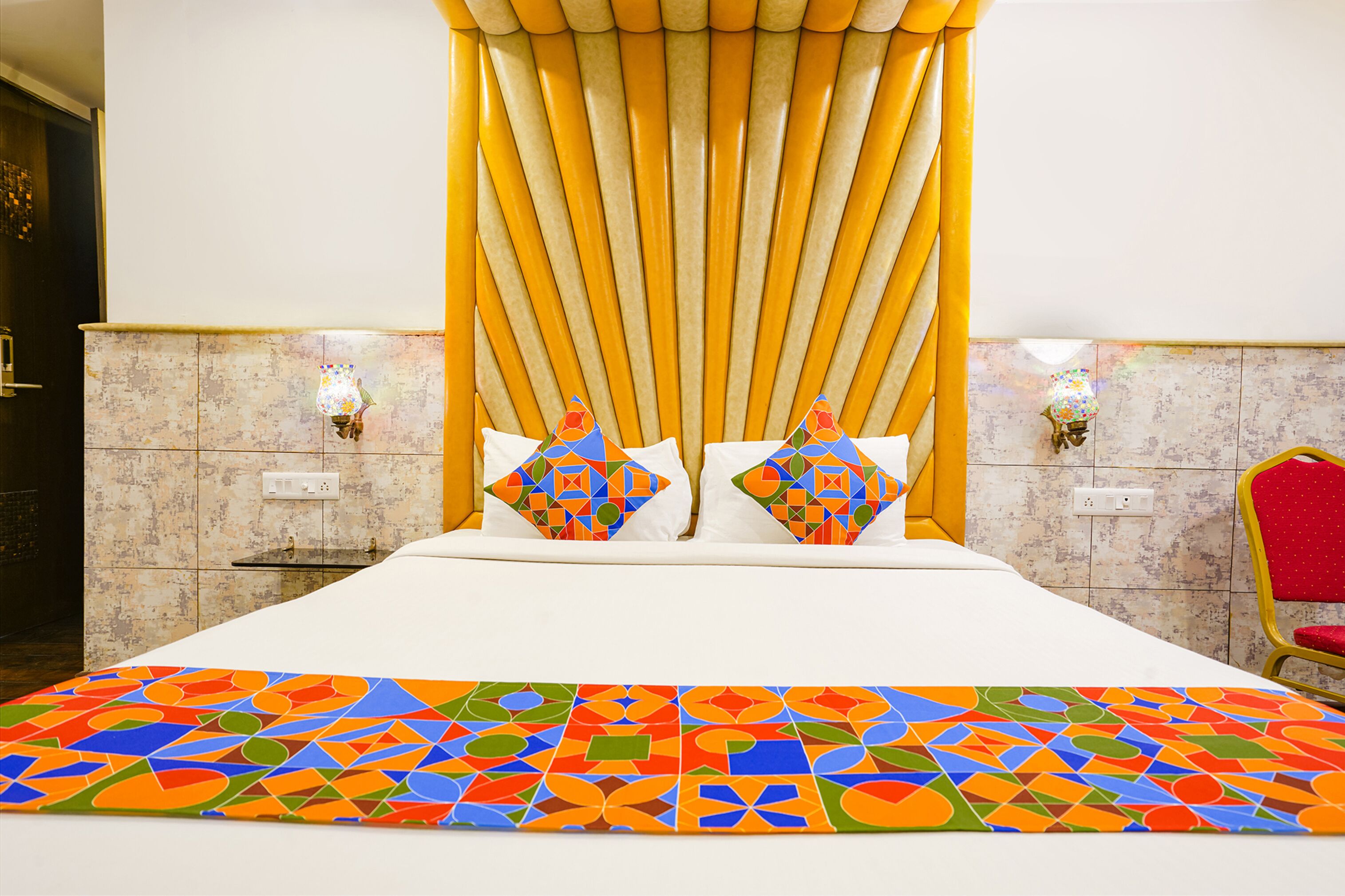 A hotel room with a large bed, a colorful geometric-patterned bedspread, two decorative pillows, a wall-mounted light fixture, and a red chair.