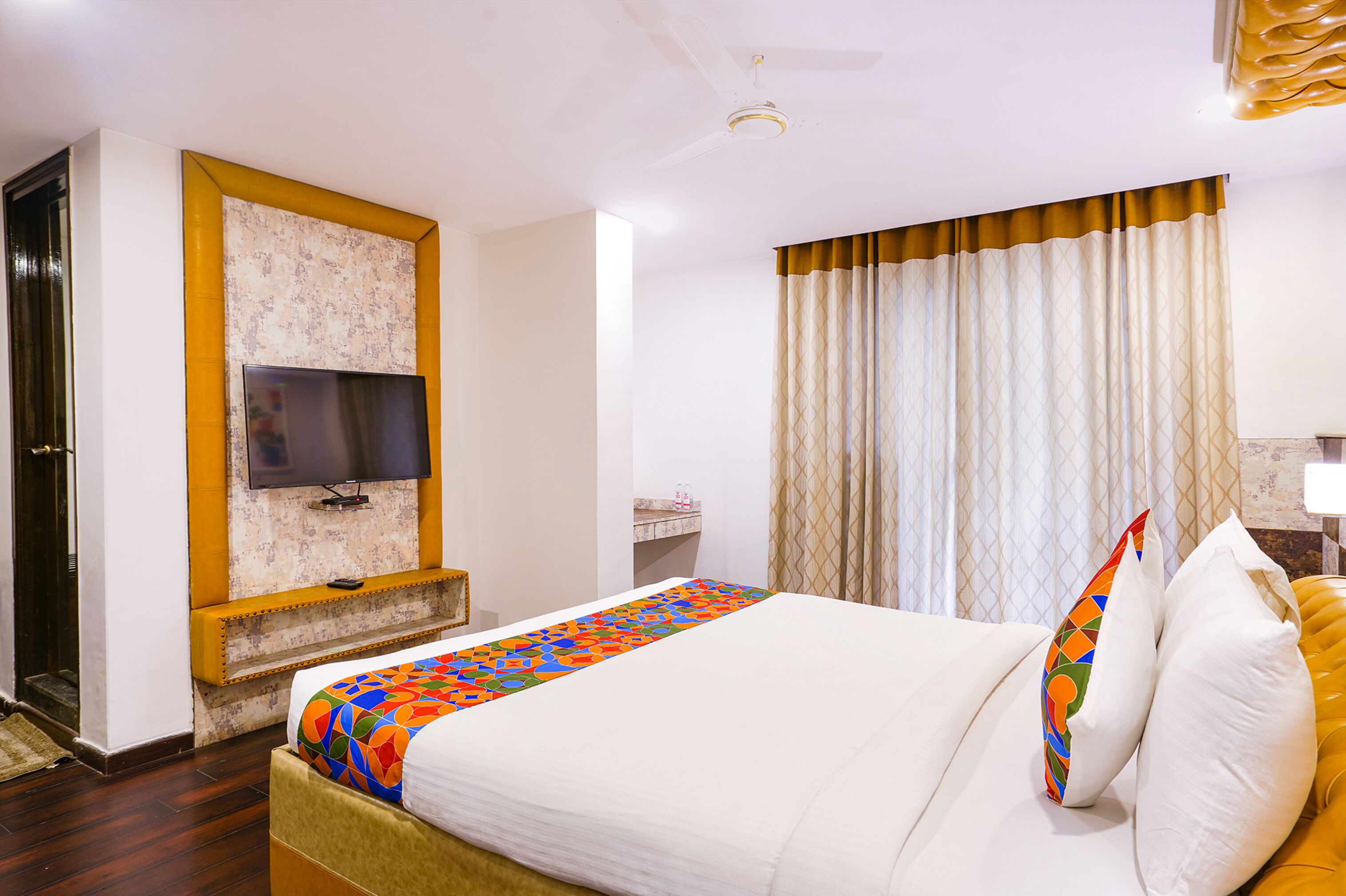 A hotel room with a bed, a television mounted on a marble wall, a bathroom with a sink, and a window with curtains.