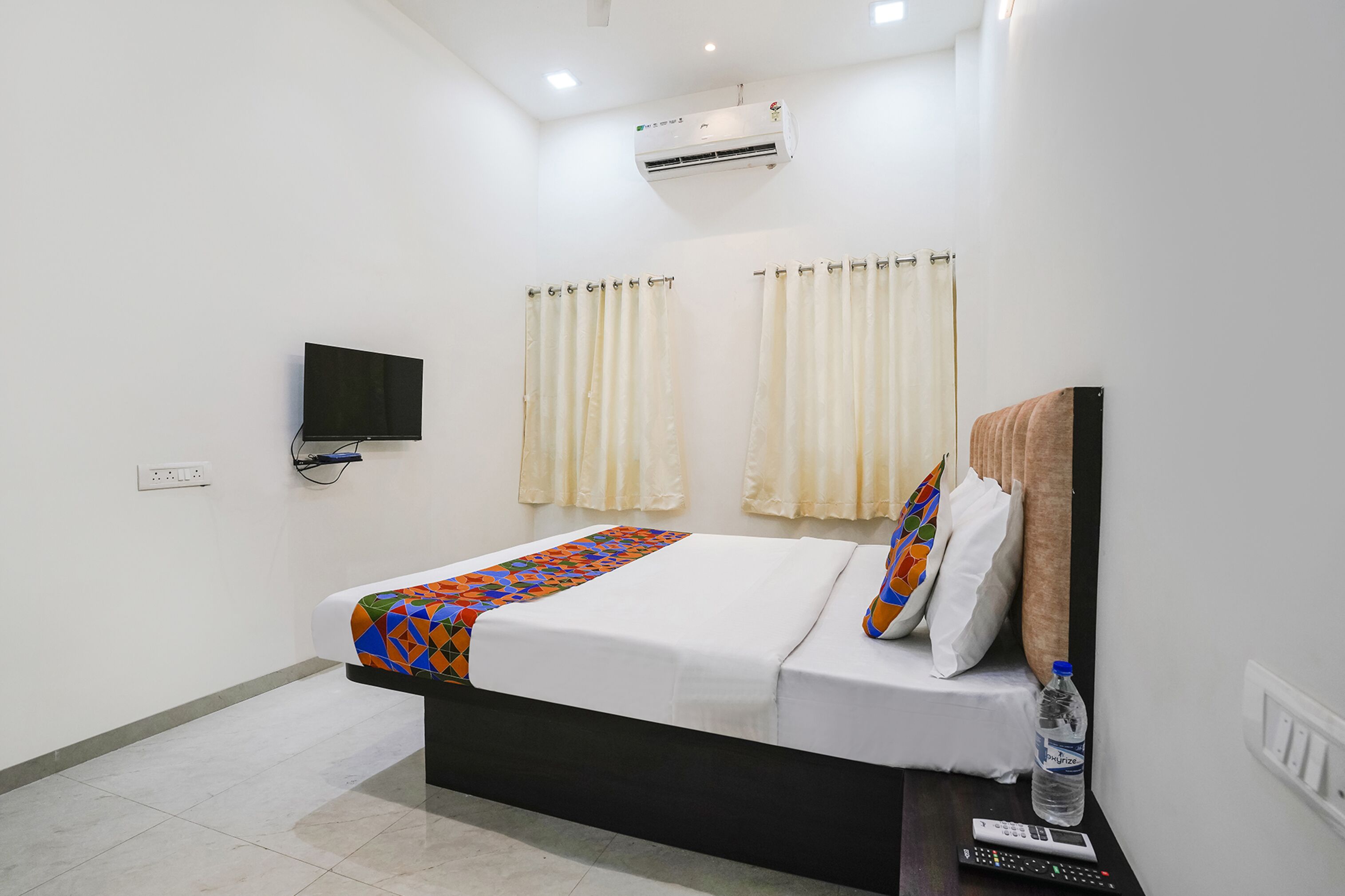 A hotel room with a bed, a TV, curtains, a pillow, a water bottle, and a remote control.