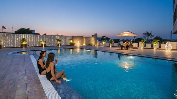 A swimming pool with two people sitting on the edge, a dining area with tables and umbrellas, and a building with string lights in the background.