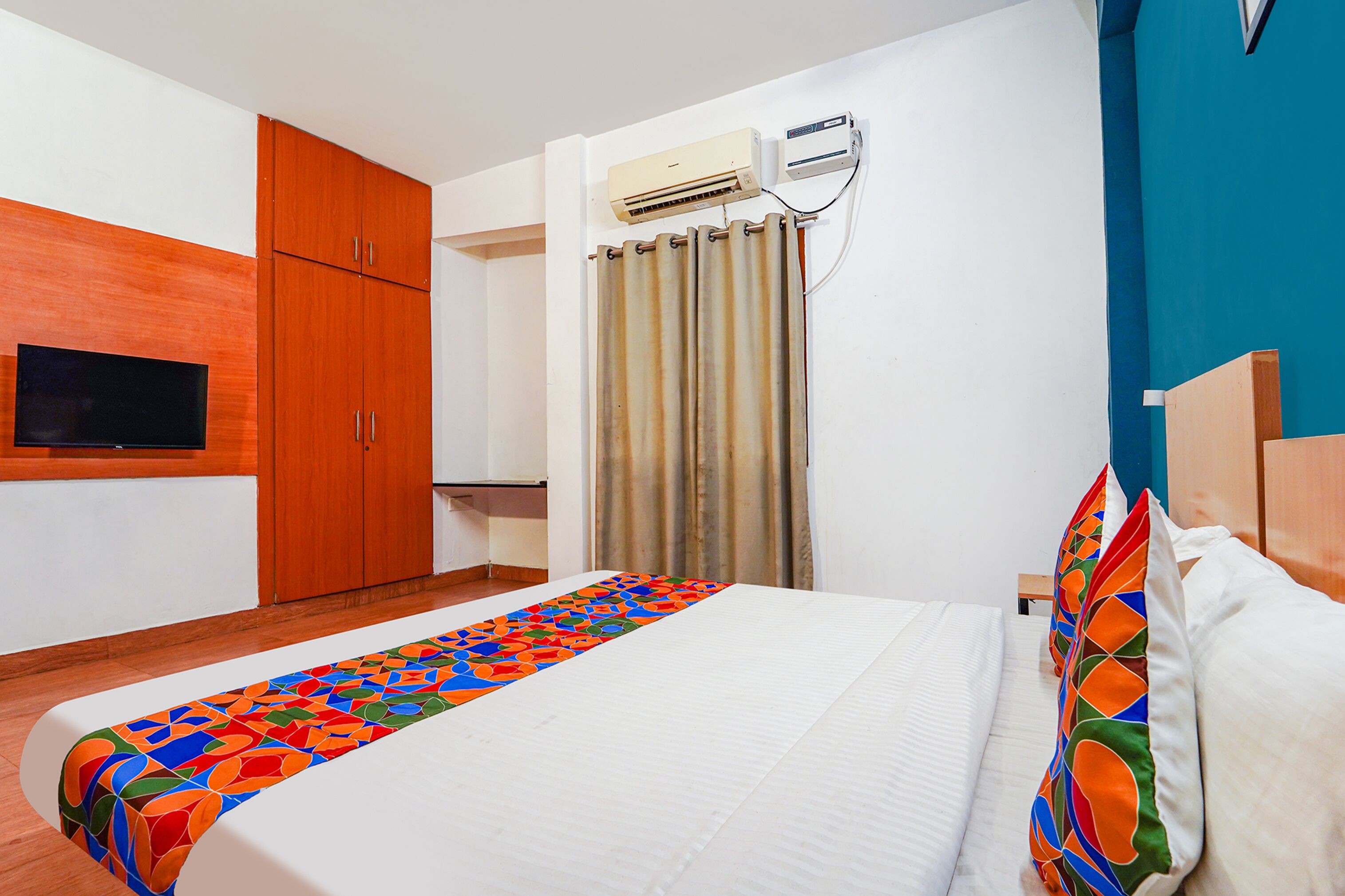 Quarto de hotel com cama, televisão, armários de madeira, ar condicionado e cortina.