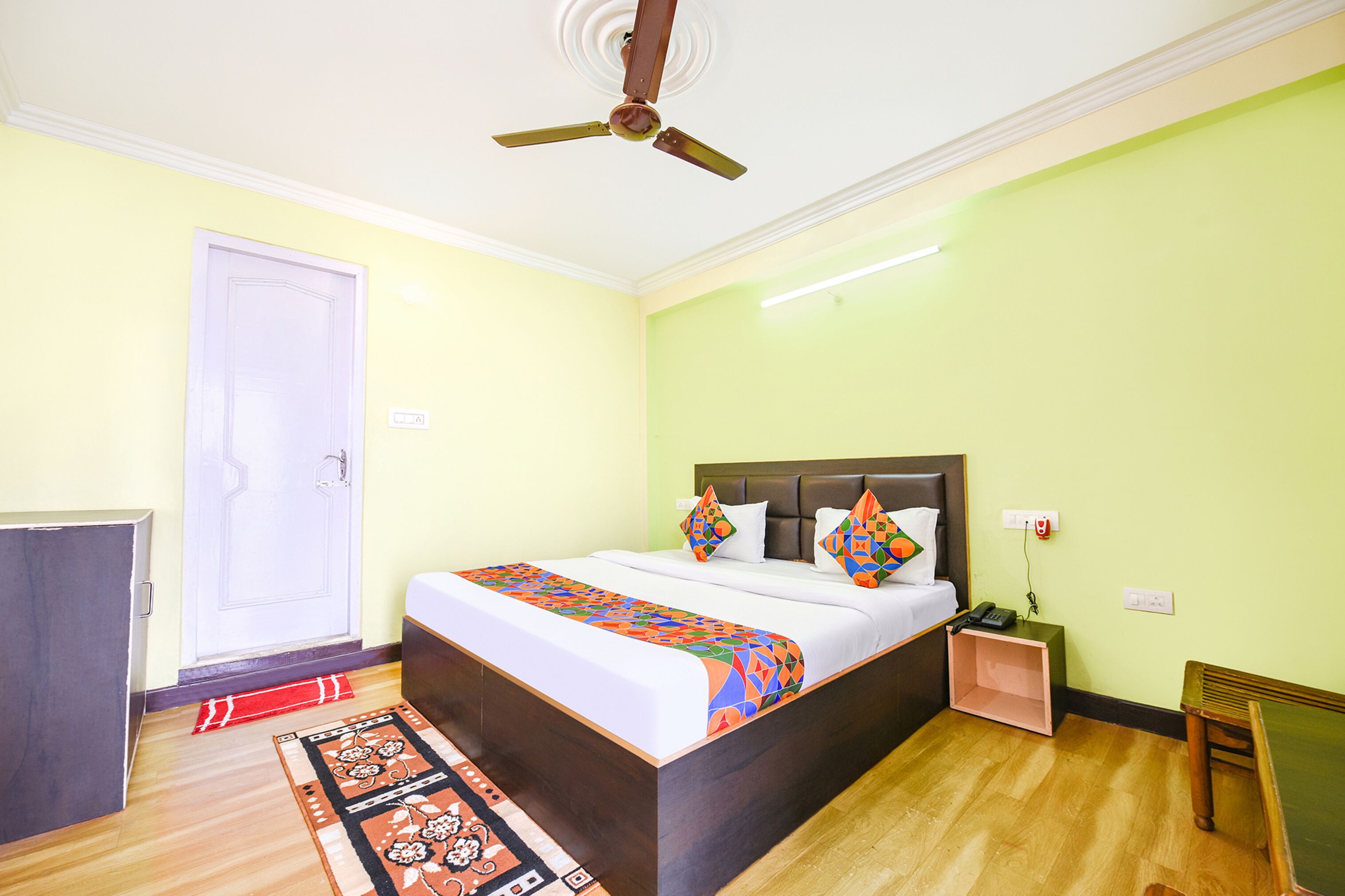 Una habitación de hotel con una cama, un ventilador de techo, una mesita de noche y otra mesita auxiliar.
