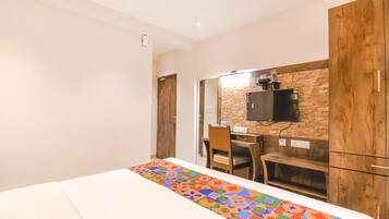 A hotel room with a bed, a desk, a chair, a TV mounted on a brick wall, and wooden cabinets.