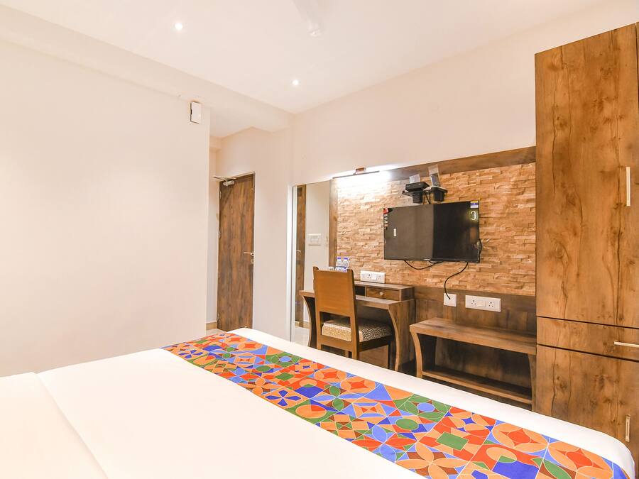 A hotel room with a bed, a desk, a chair, a TV mounted on a brick wall, and wooden cabinets.