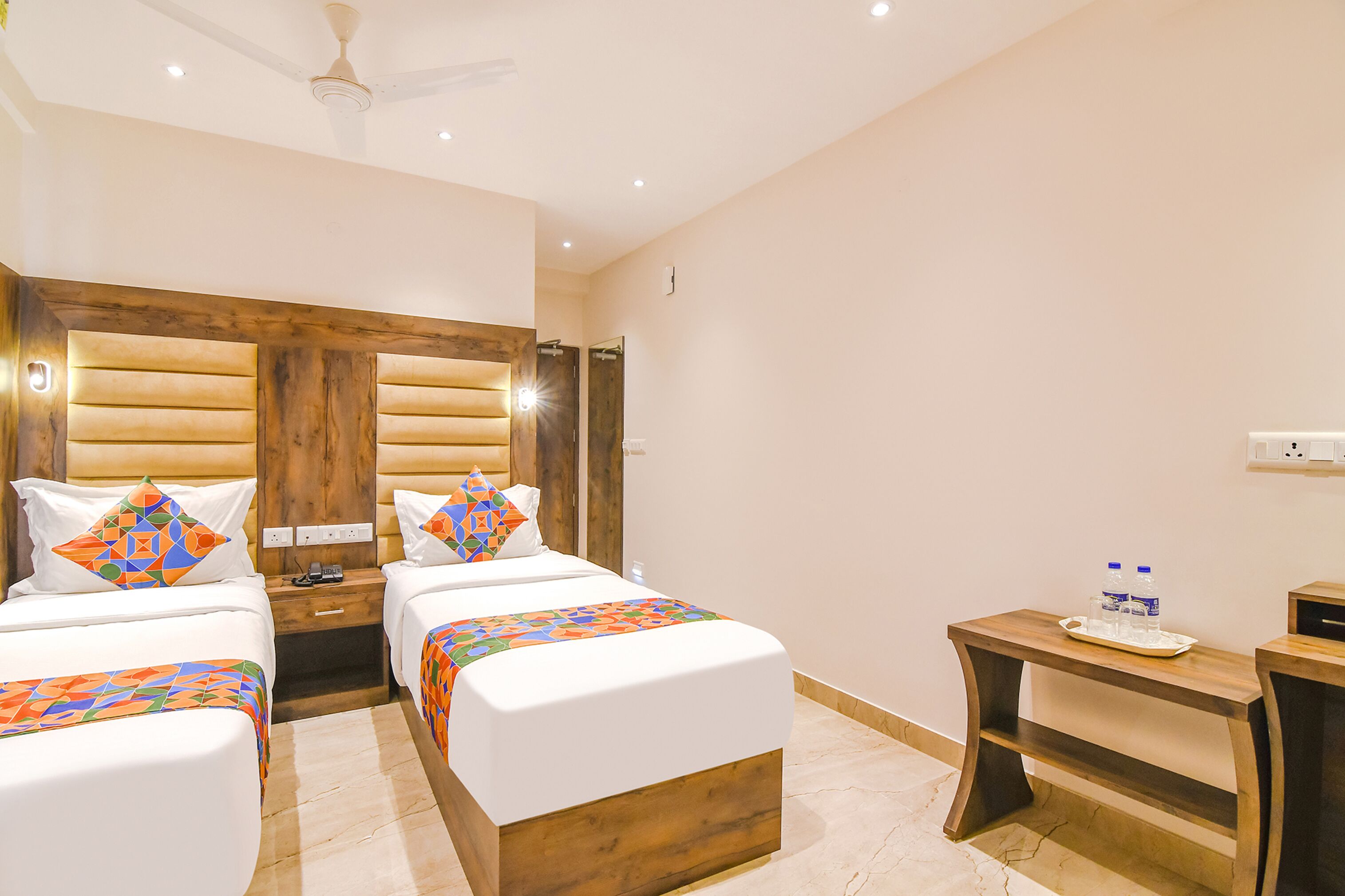 Habitación de hotel con dos camas, cabecera de madera, una mesita con botellas de agua y un ventilador de techo.