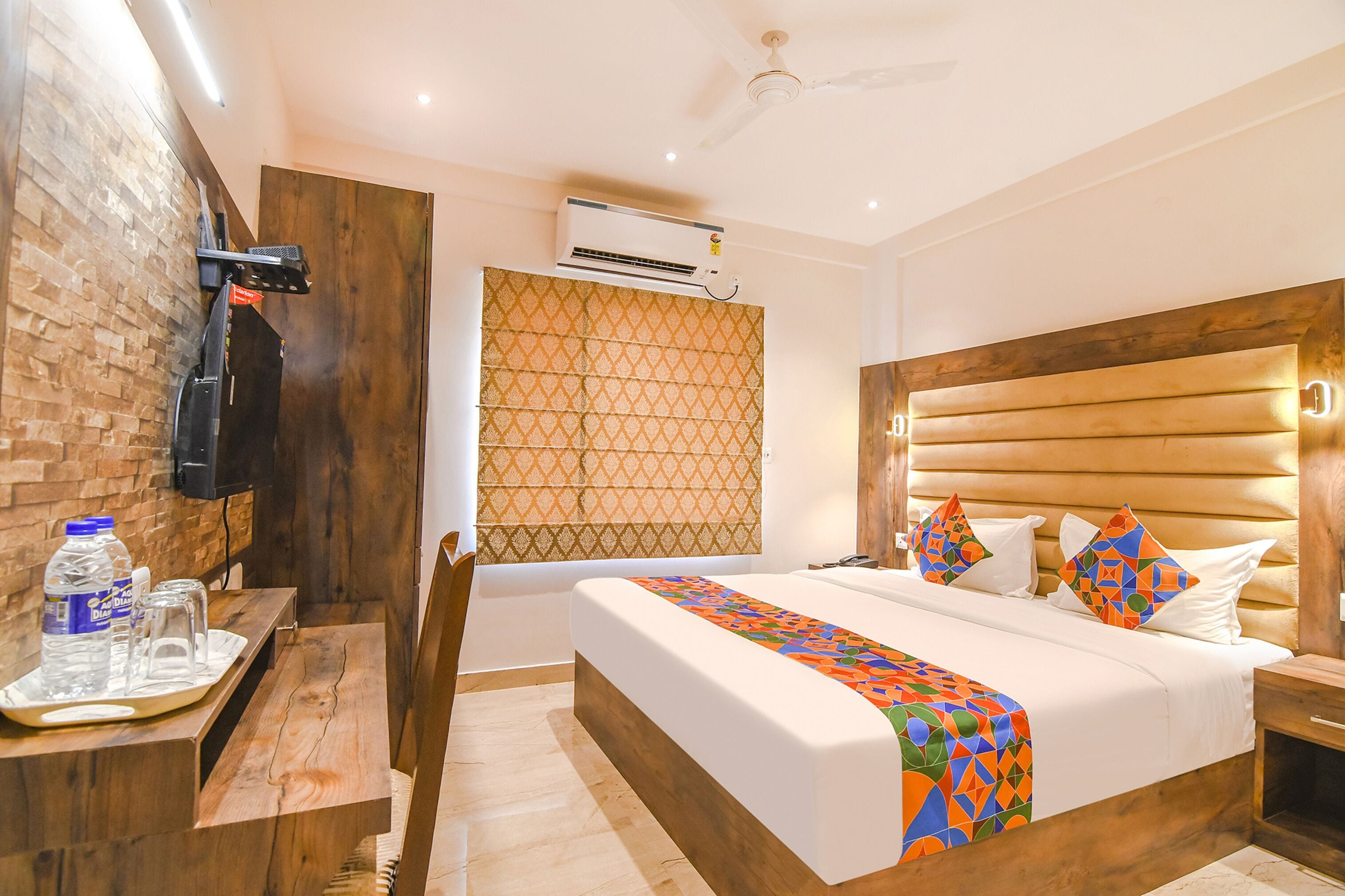 Una habitación de hotel con cabecera de madera, cama con ropa de cama colorida, escritorio con bandeja y aire acondicionado de pared.
