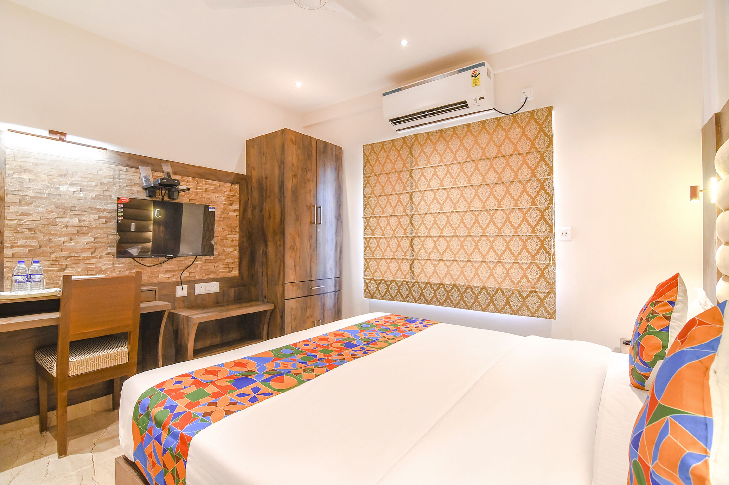 Una habitación de hotel con cama, un escritorio con silla, un televisor montado en una pared de ladrillo y una cortina estampada.