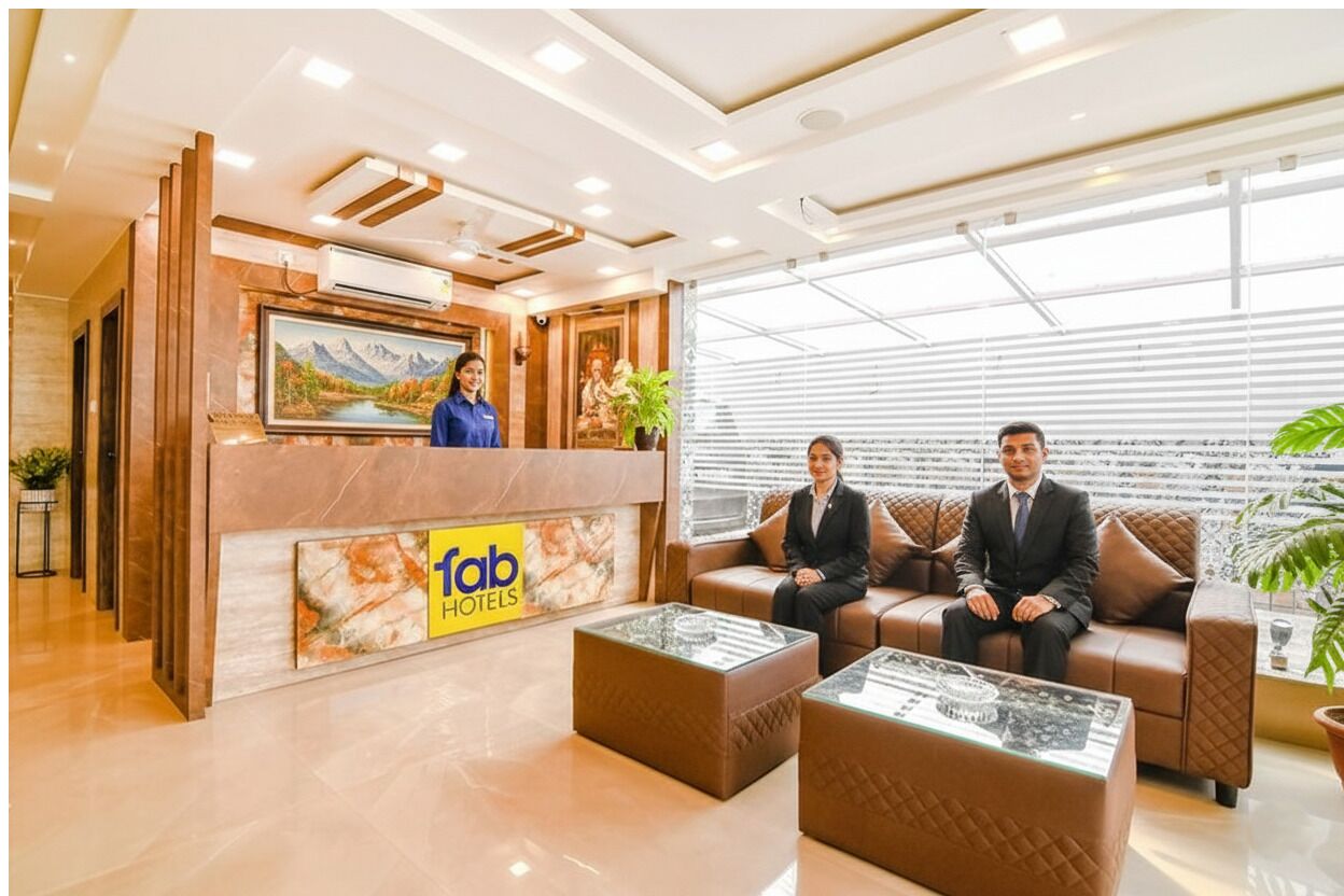 Un espace d’accueil d’hôtel comprenant un comptoir de réception, deux clients assis et une grande fenêtre avec des stores.
