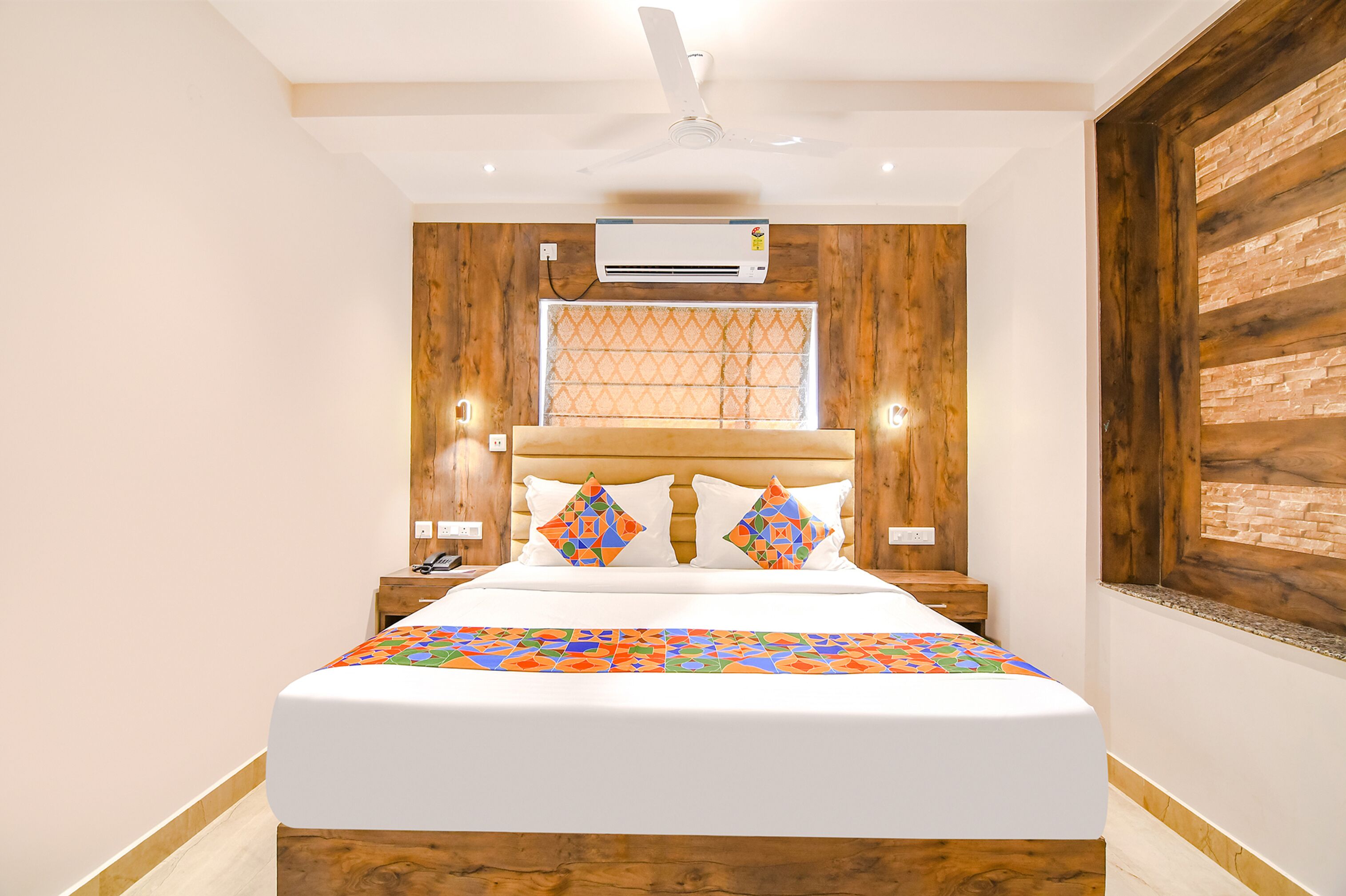 Een slaapkamer met een houten hoofdbord, een bed met kleurrijke kussens, een airconditioning en een raam met een decoratief blind.