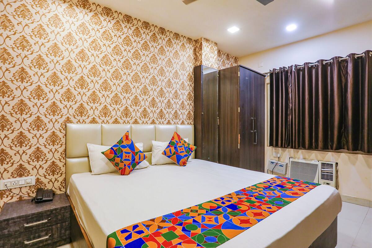 A hotel room with a patterned wallpaper, a bed with a colorful geometric bedspread, a wardrobe, and a window with curtains.