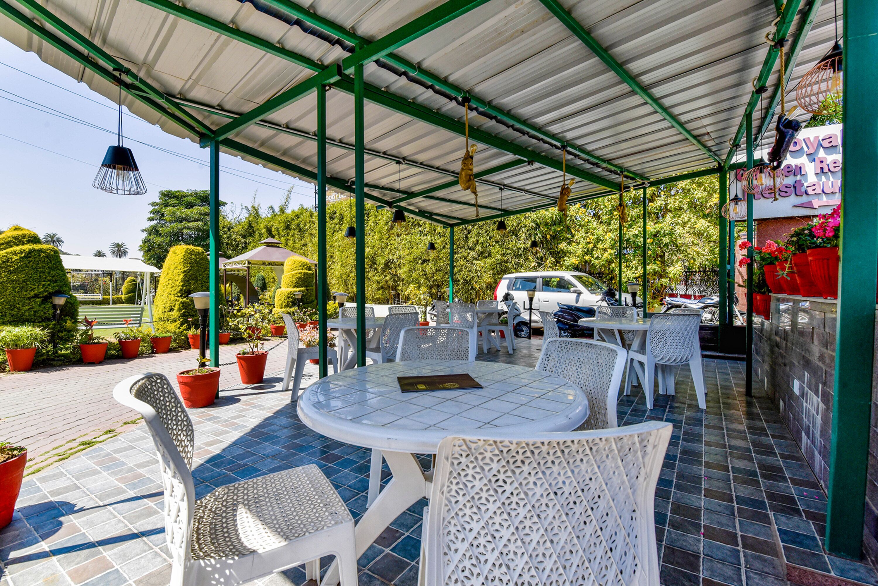An outdoor seating area with round tables and white chairs, surrounded by potted plants and a covered structure.