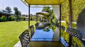 A covered outdoor dining area with a glass table and chairs, surrounded by greenery and a building in the background. - Fabhotel Resort Royal Garden Retreat (Abu Road)