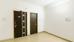 A room with two doors, each with a metal handle and a keyhole. The walls are plain white, and there is a light switch on the wall. - FabHotel The Hudco Grand (Patna)