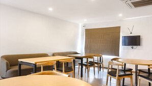 A modern dining area with wooden tables and chairs, a sofa, and a flat-screen TV mounted on the wall. - FabExpress Vivek (Prayagraj)