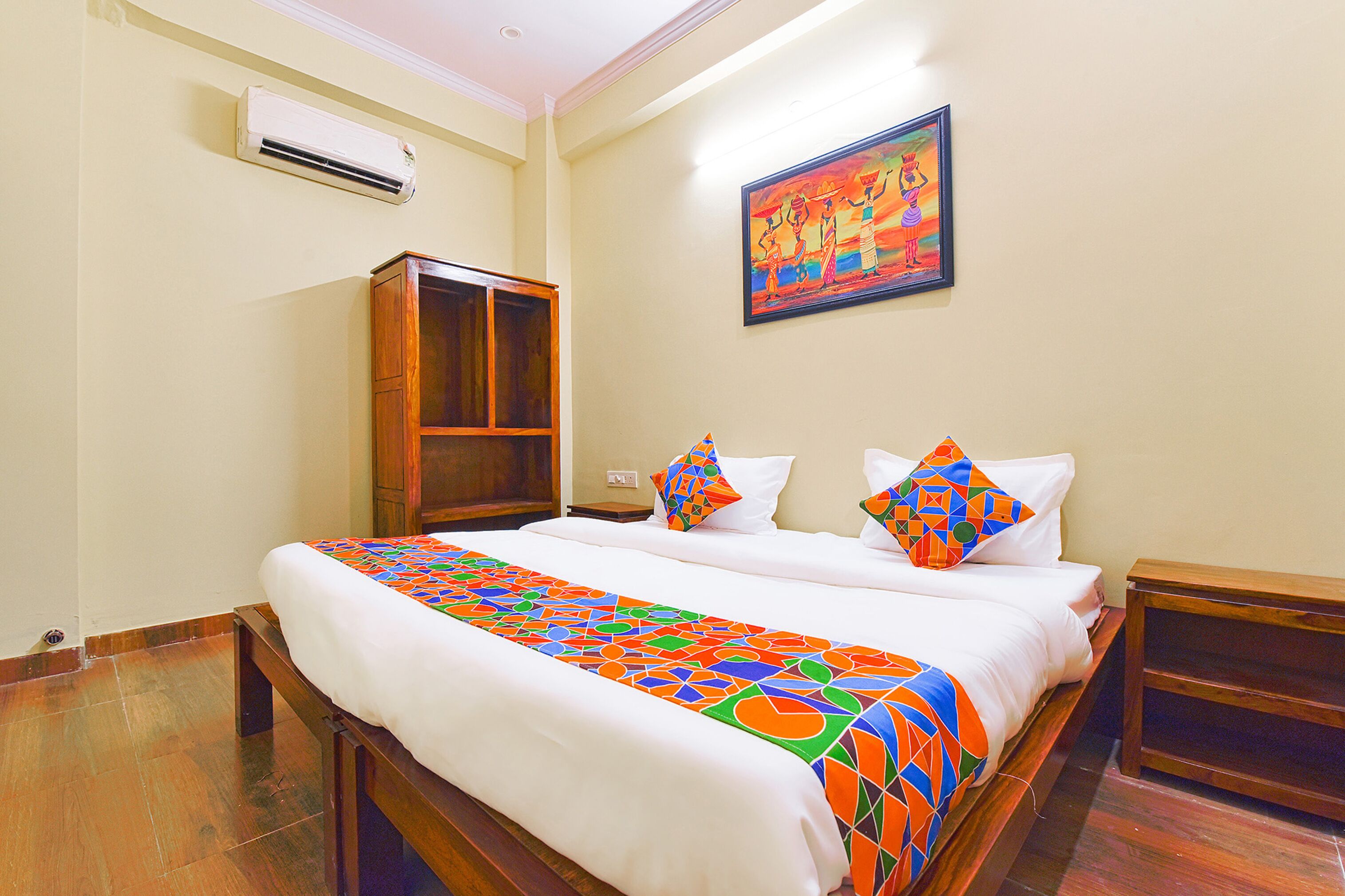 A hotel room with a large bed, a wooden wardrobe, a colorful artwork on the wall, and an air conditioning unit.
