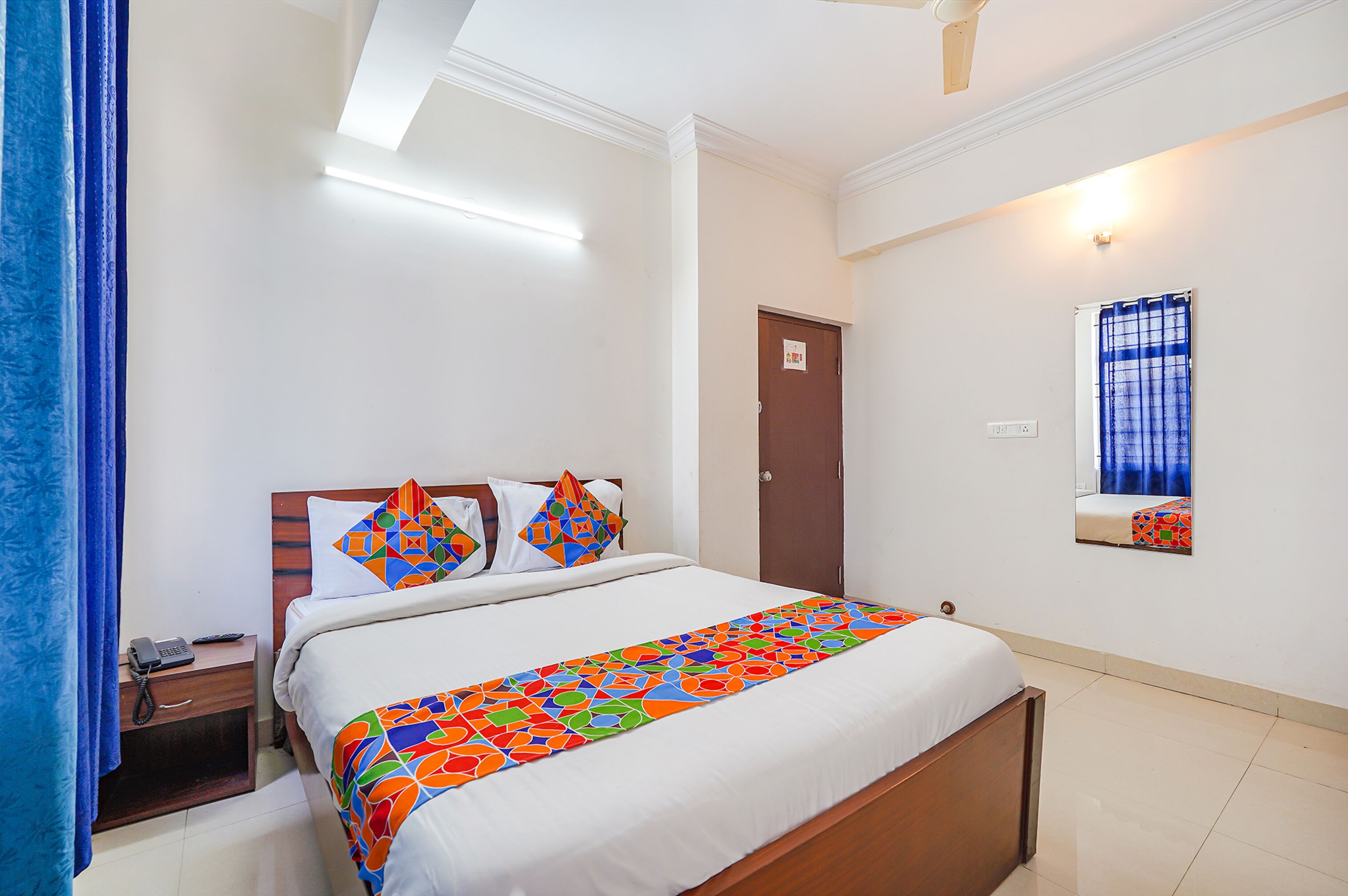A hotel room with a bed, a nightstand with a phone, a door, and a window with a curtain.