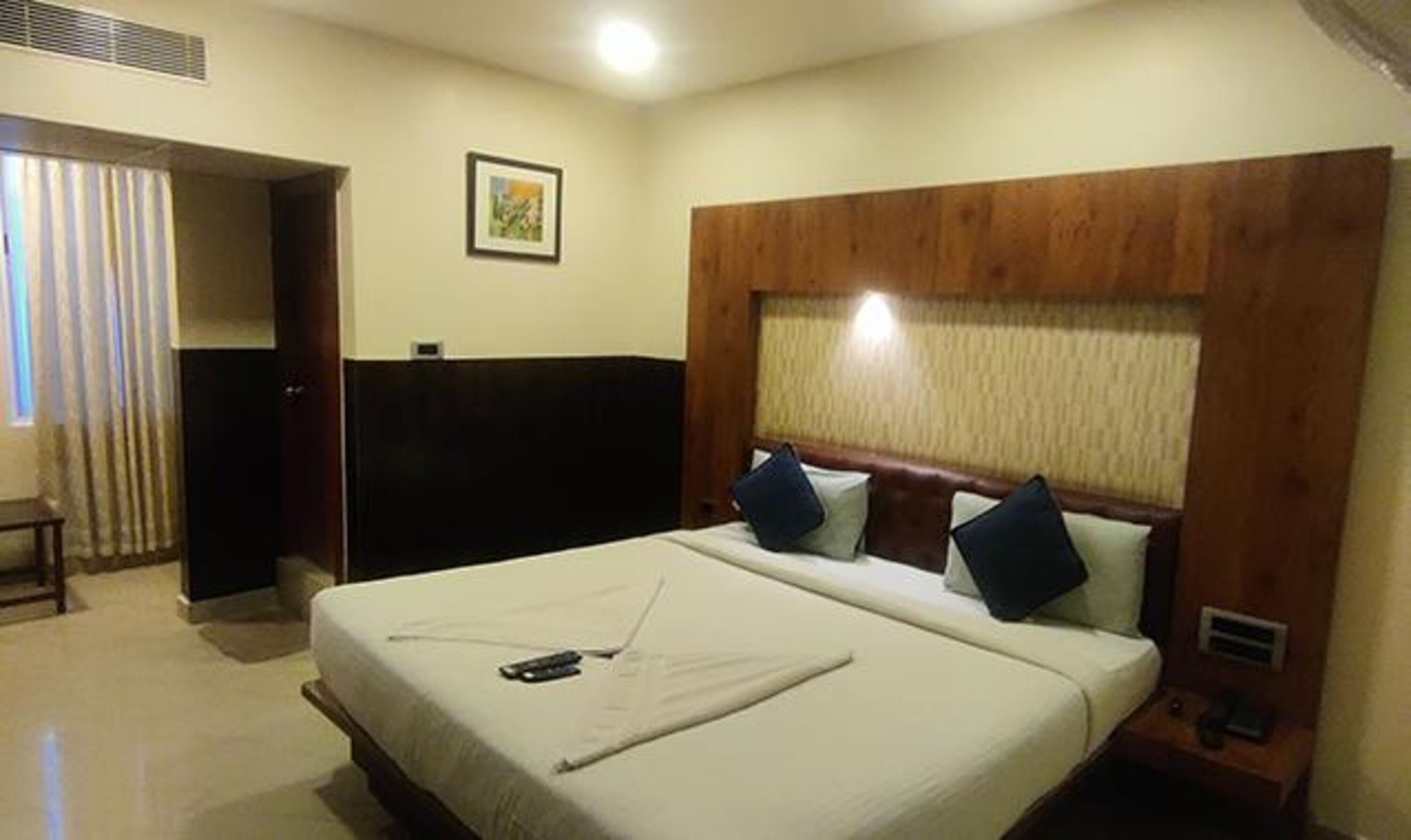 A hotel room with a large bed, a wooden headboard, a bedside table, and a window with curtains.
