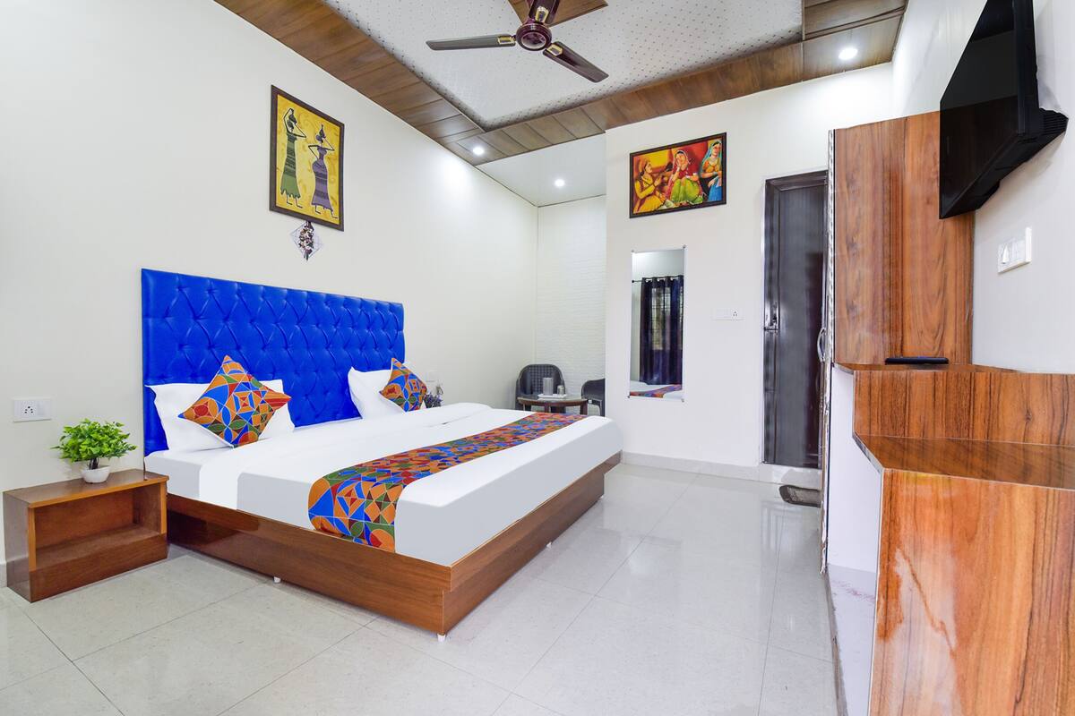 A modern bedroom with a tufted blue headboard, a bed with colorful geometric bedding, a wooden nightstand with a plant, a ceiling fan, and a wooden wardrobe.