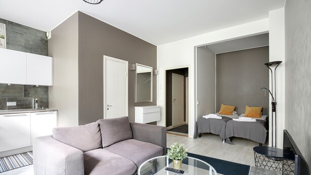 A modern living room with a sofa, a coffee table, and a TV. There is a kitchen area with cabinets and a countertop. - Kotimaailma Aparthotel Kamppi - Studios (Helsinki)