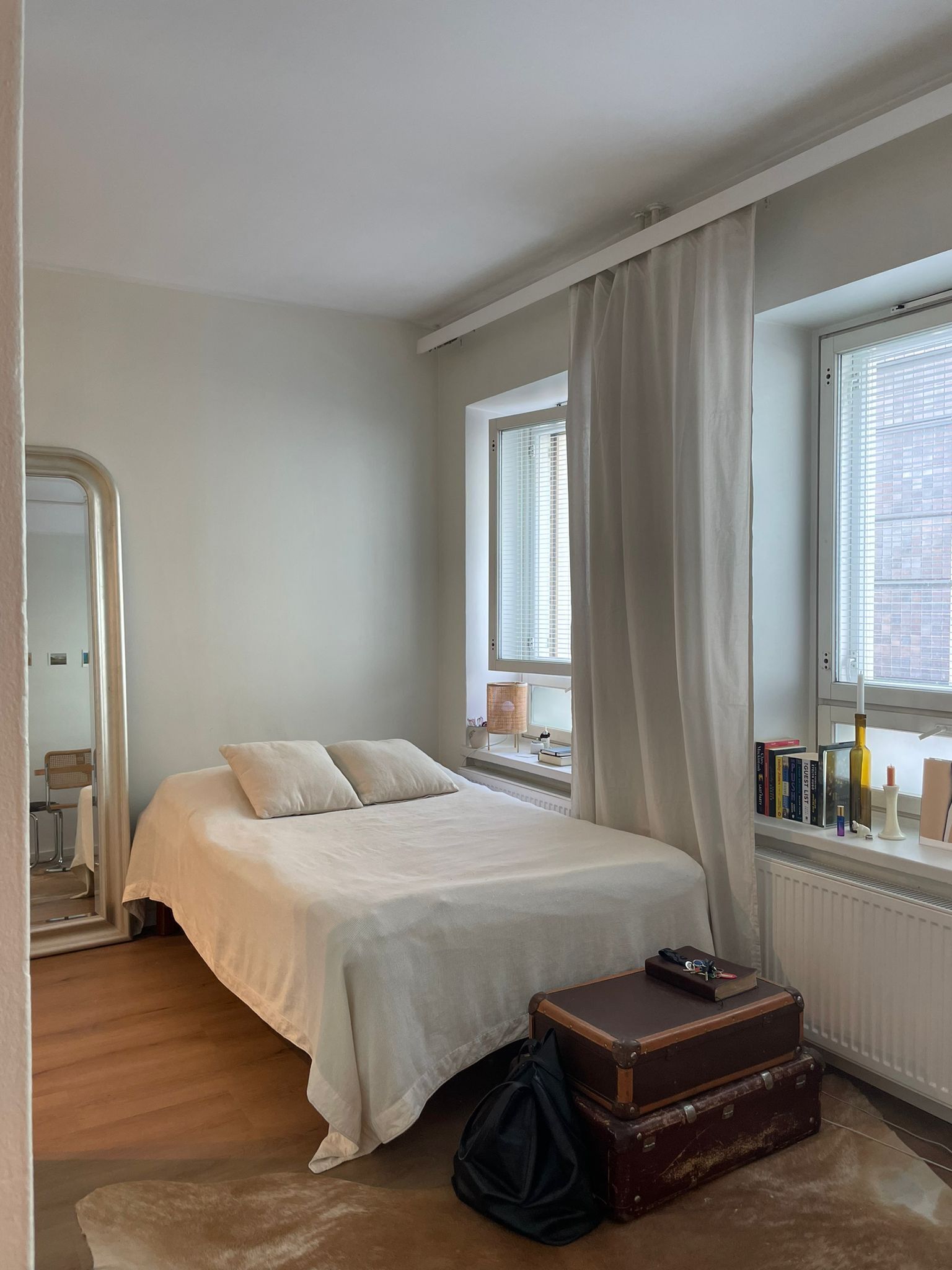 A bedroom with a bed, a window with blinds, a radiator, a suitcase, and a mirror.