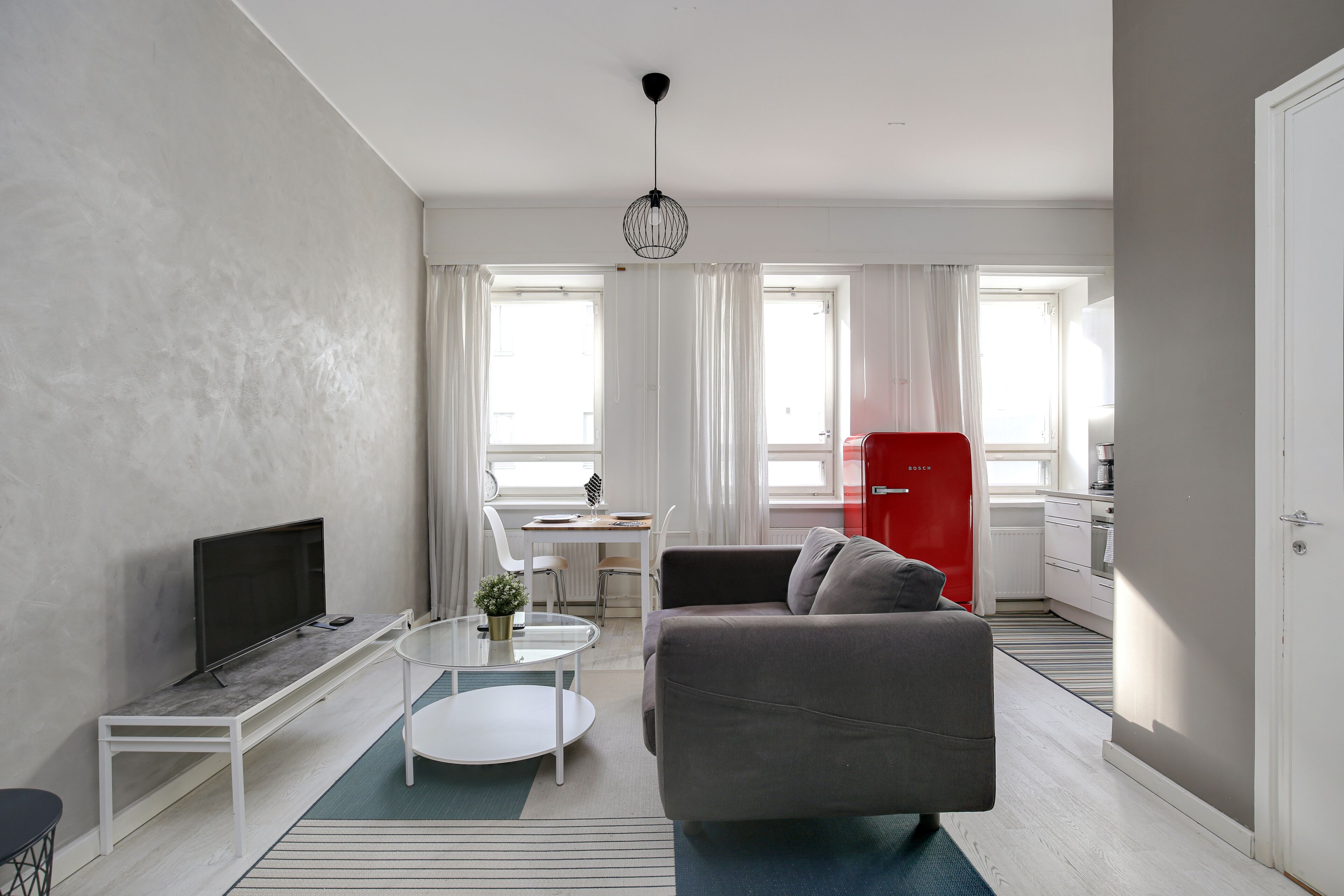 Ein modernes Wohnzimmer mit einem grauen Sofa, einem Fernsehgerät auf einem weißen Standfuß, einem kleinen Tisch mit einer Pflanze und einem roten Kühlschrank im Hintergrund.
