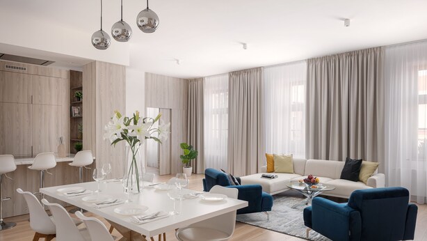 A modern living room with a dining area, featuring a white dining table, blue sofas, and a large window with curtains.