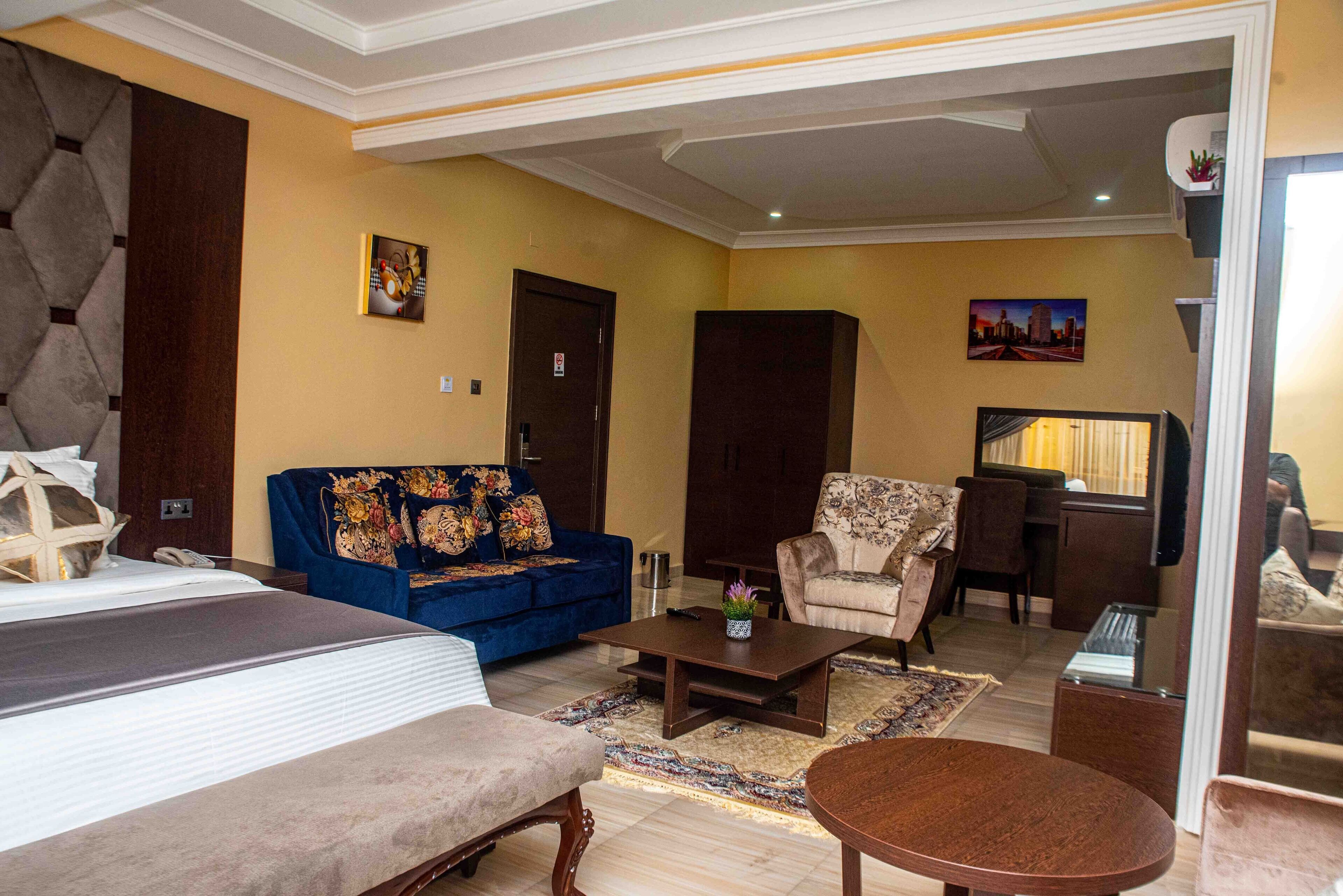 Habitación de hotel con una cama, un sofá azul, un sillón color beige, una mesa de centro de madera y una mesita con un florero.
