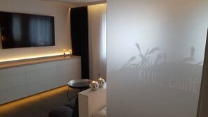 A modern bathroom with a large mirror, a flat-screen TV mounted on the wall, and a frosted glass partition with a palm tree design. - Kleine Düne (Cuxhaven)