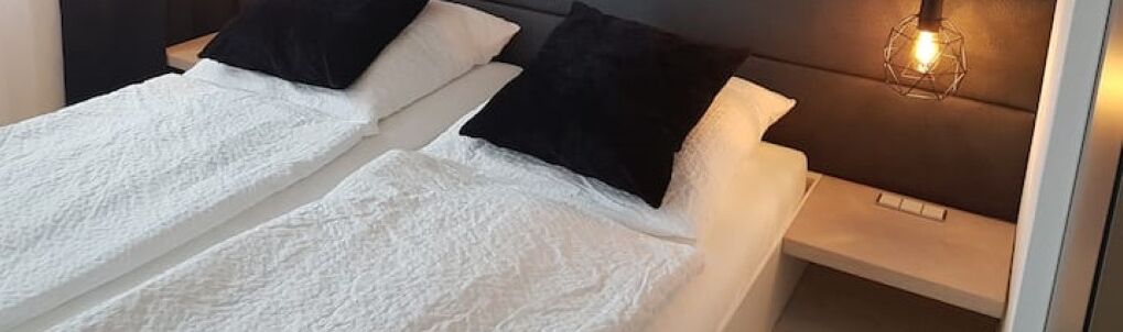 A double bed with white linens and black pillows, a bedside table, and a wall-mounted light fixture.